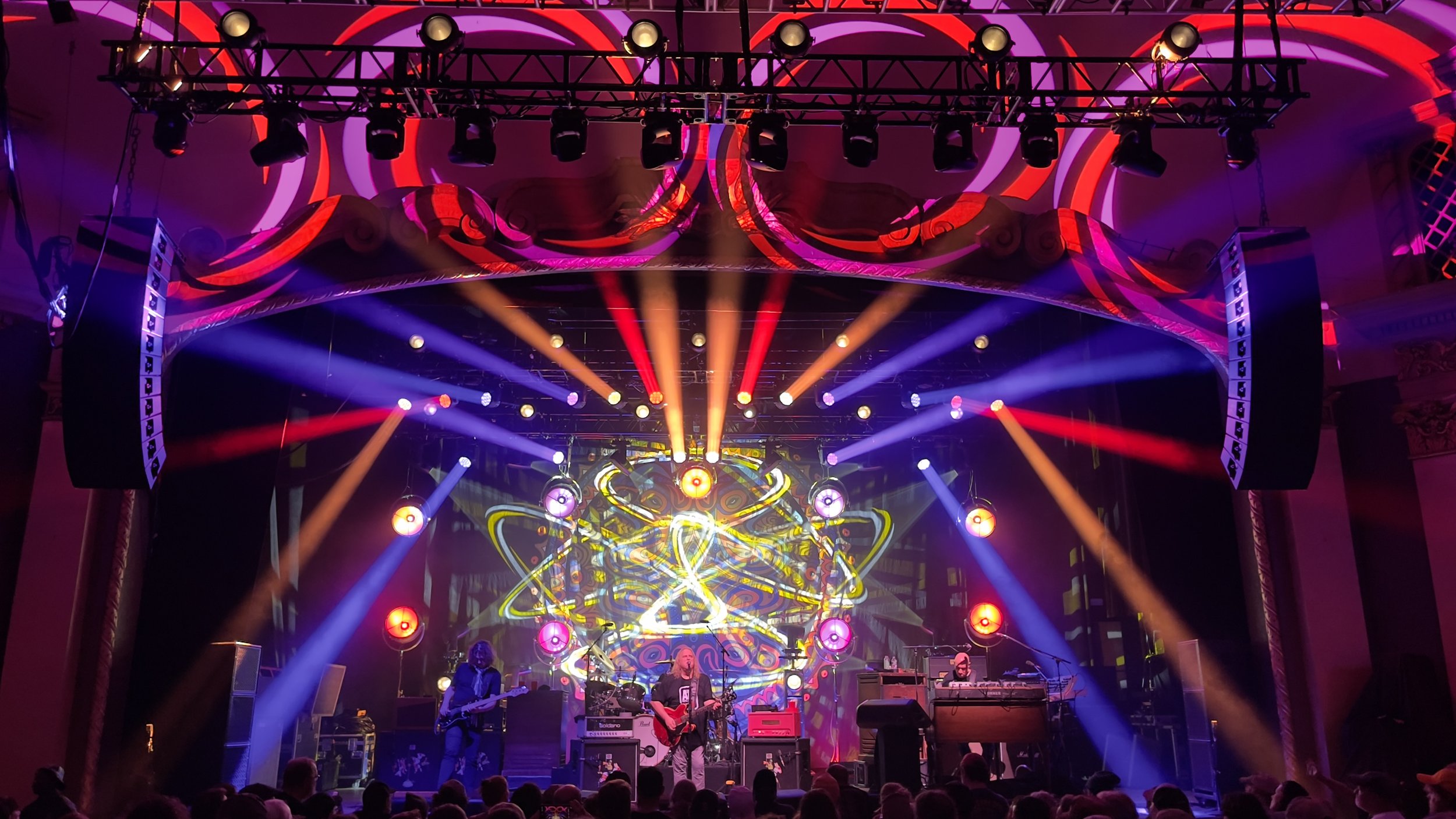 A colorful concert stage with bright lights and psychedelic background, featuring musicians performing and an audience in front.