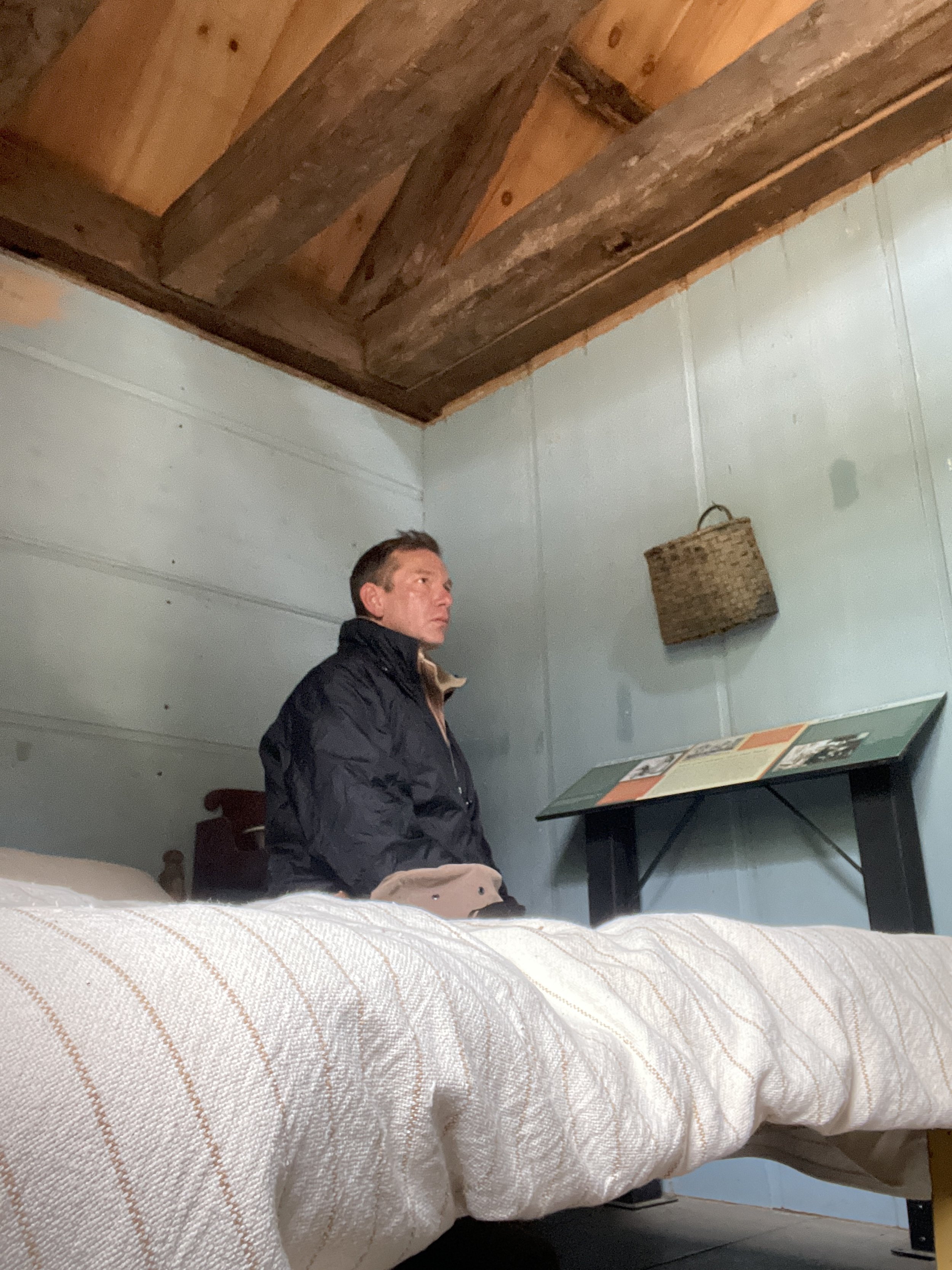 A man sitting on a bed inside a rustic room with light green walls and a wooden ceiling. He is wearing a black jacket and looking to the left. There is a small table with an informational display and a woven basket hanging on the wall.