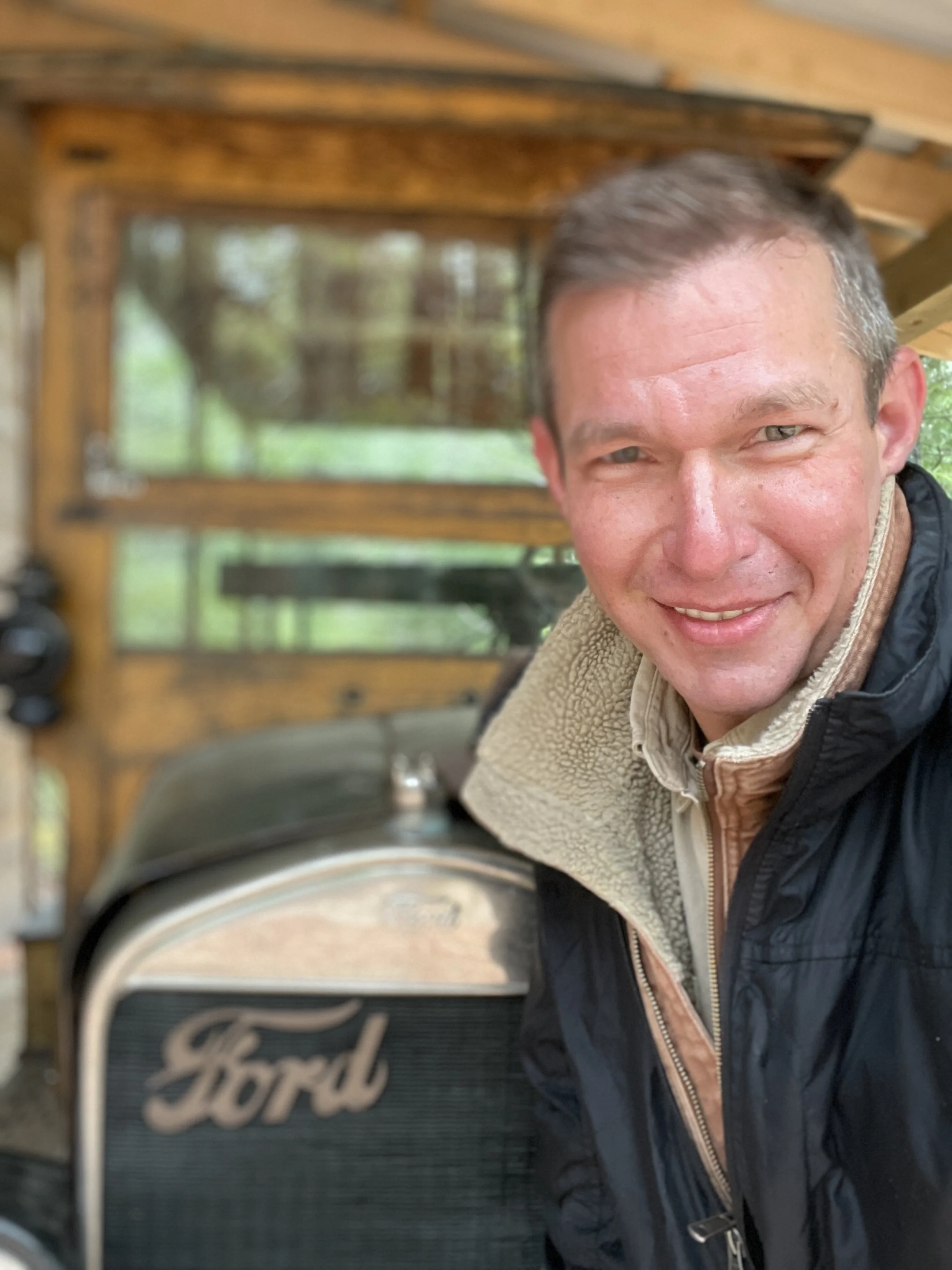 A man smiling for a selfie in front of a vintage Ford car inside a wooden shed or garage.