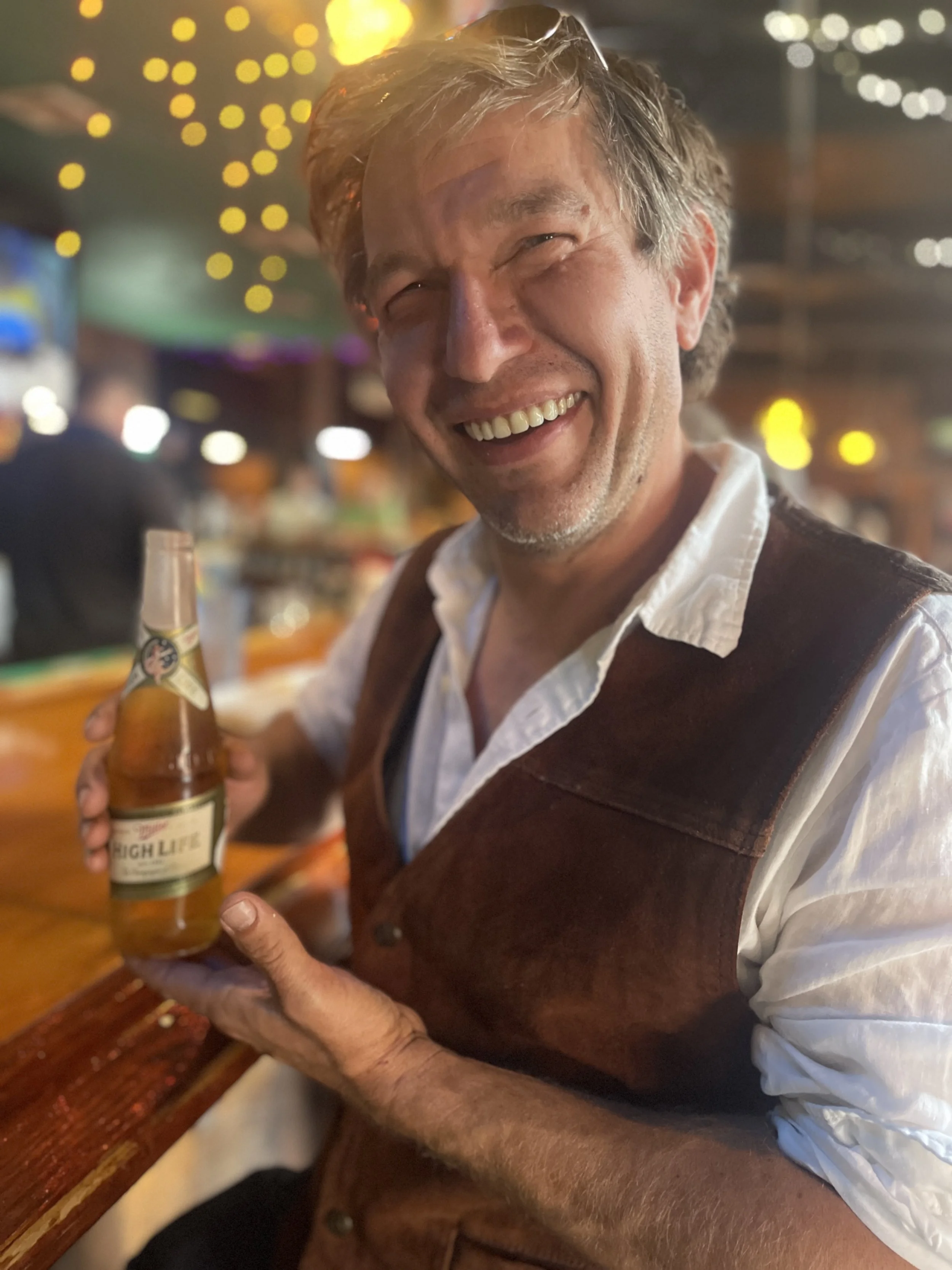 A smiling man, named Julian Schlaver, holding a beer bottle in a lively bar or restaurant with warm lighting and blurred background.