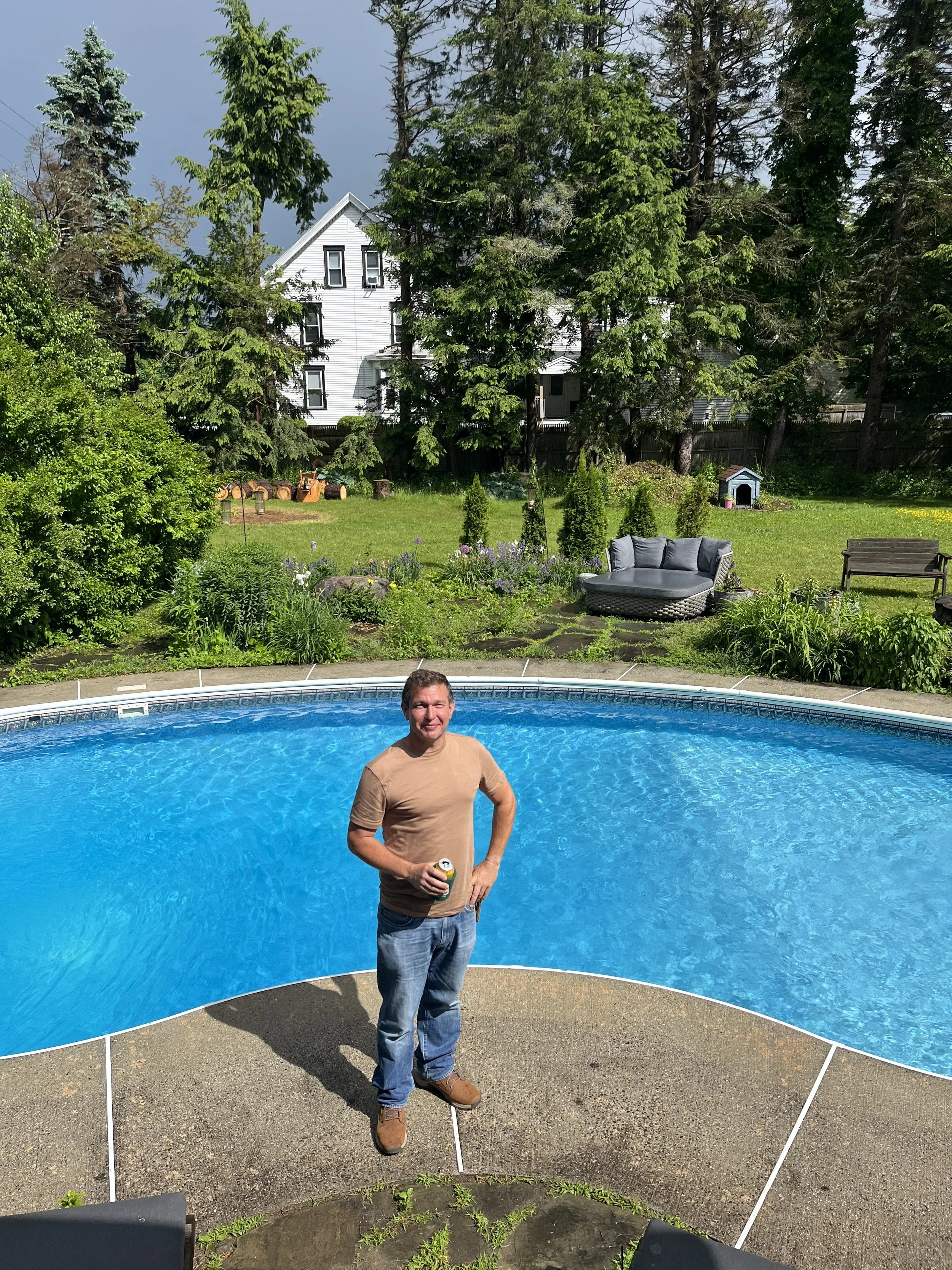 Man, named Julian Schlaver, standing by a pool holding a can, smiling. Green garden with trees and outdoor furniture in the background.