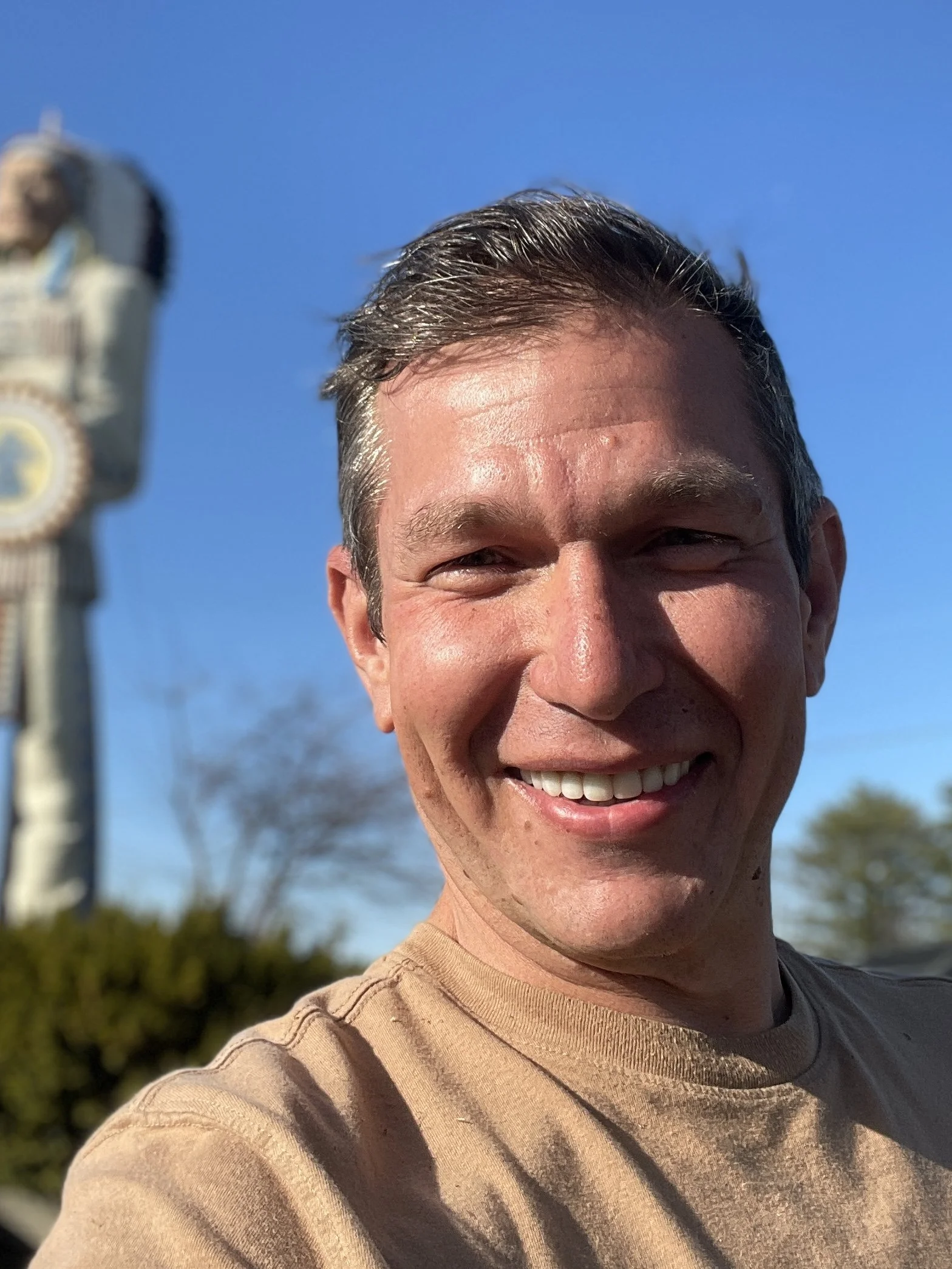 A smiling man, named Julian Schlaver, with short dark hair takes a selfie outdoors under a clear blue sky, with a blurred monument or tall structure in the background.