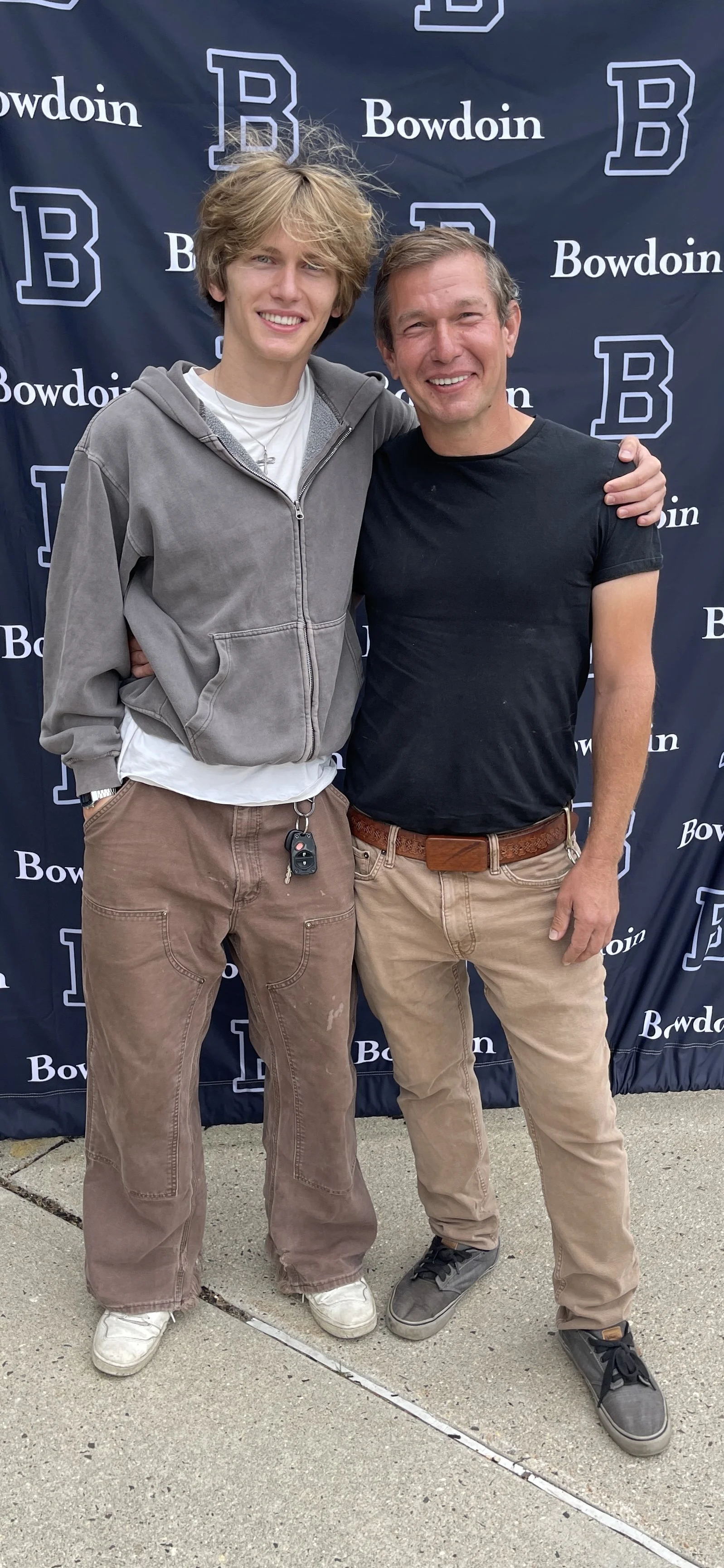 Two men standing together outdoors, hugging, in front of a backdrop with the word 'Bowdoin' and a large letter 'B' logo, smiling at the camera.
