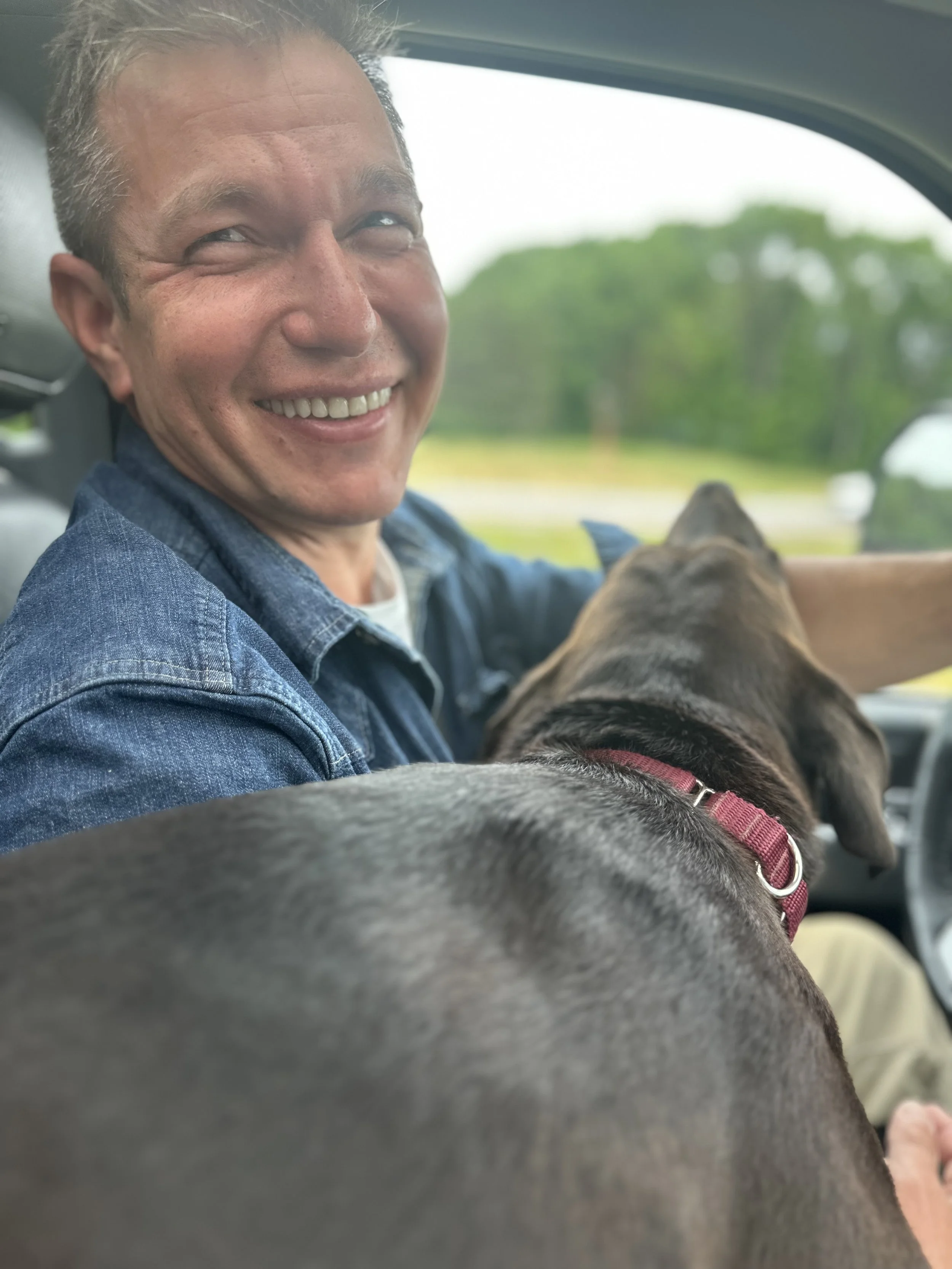 A smiling man, named Julian Schlaver, driving a vehicle with a brown dog sitting on his lap, facing forward, while the man looks back and smiles.