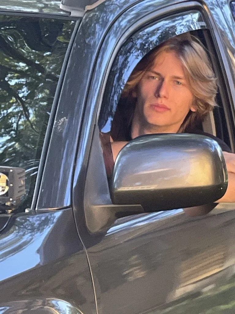 A young man with long blond hair sitting in the driver's seat of a black vehicle, looking out the window with a serious expression.