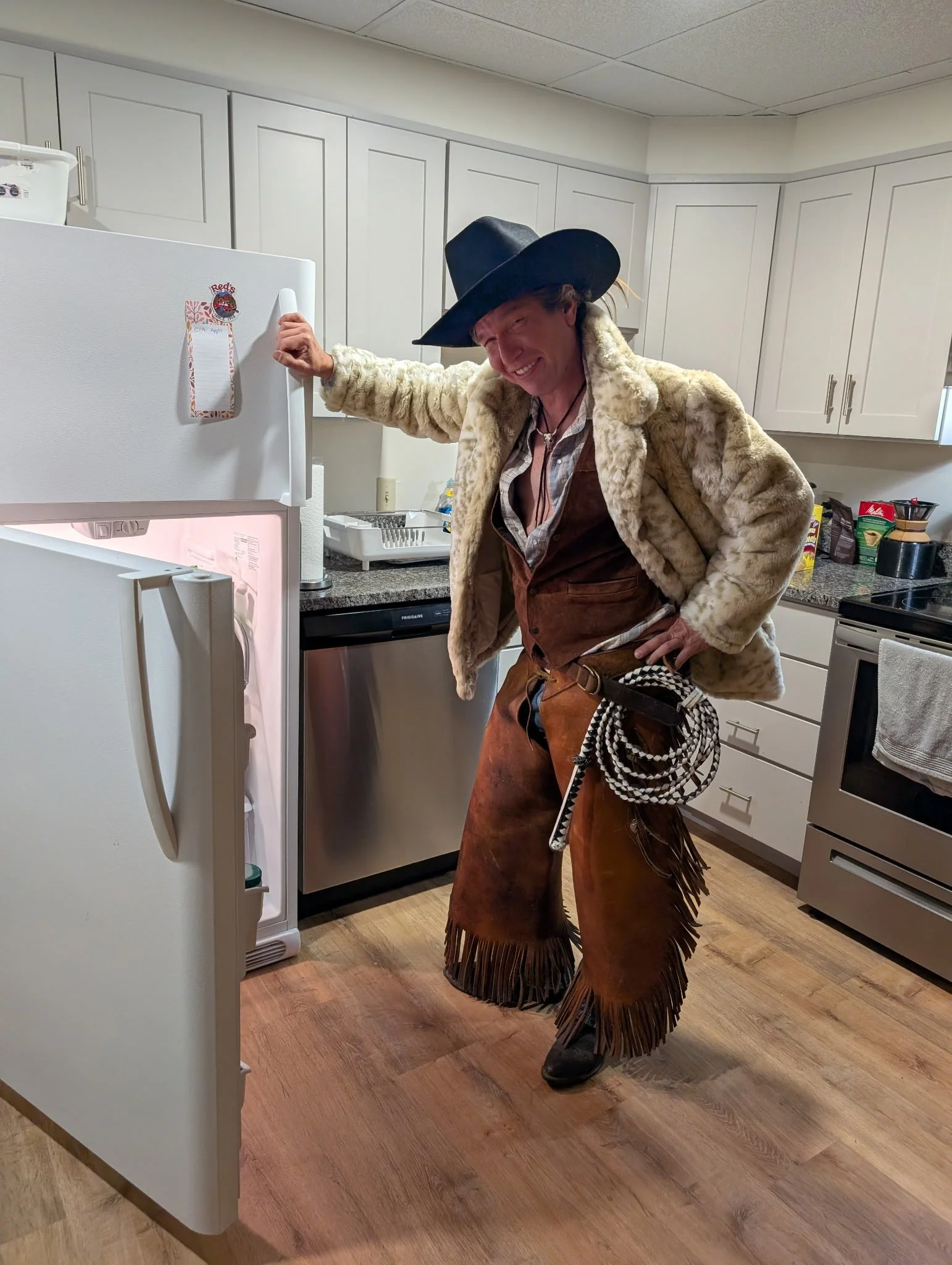 Person dressed as a cowboy opening a refrigerator in a kitchen, wearing a black cowboy hat, a fluffy fur coat, a western-style vest, fringed pants, and boots.