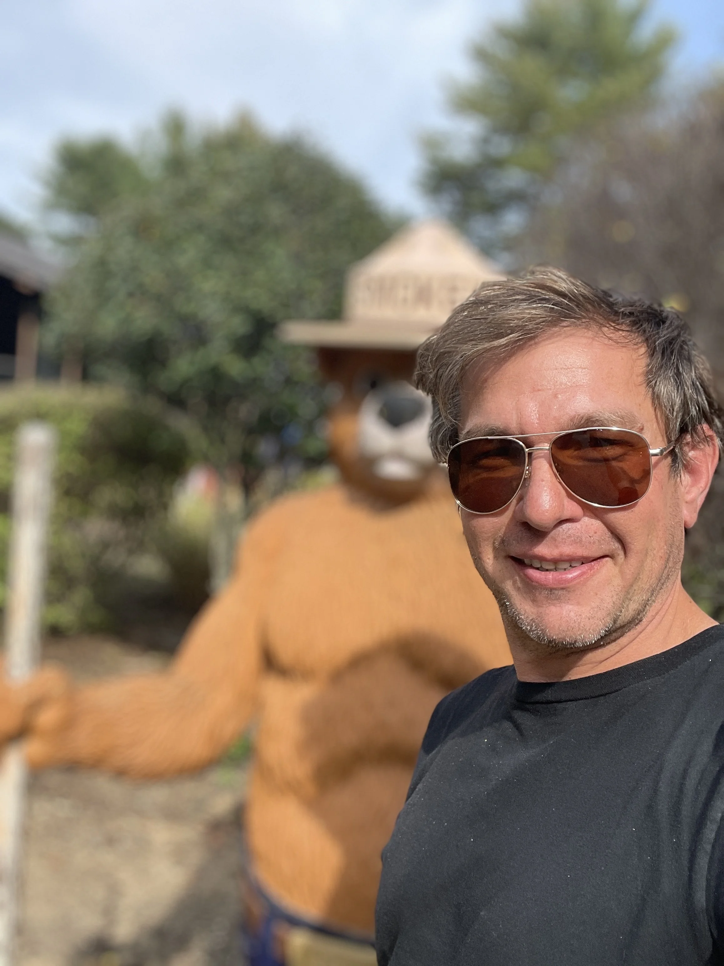 A man, named Julian Schlaver, wearing sunglasses smiling in front of a large statue of a bear with a hat and a sign that says "SILVER" in the background, outdoors on a sunny day.