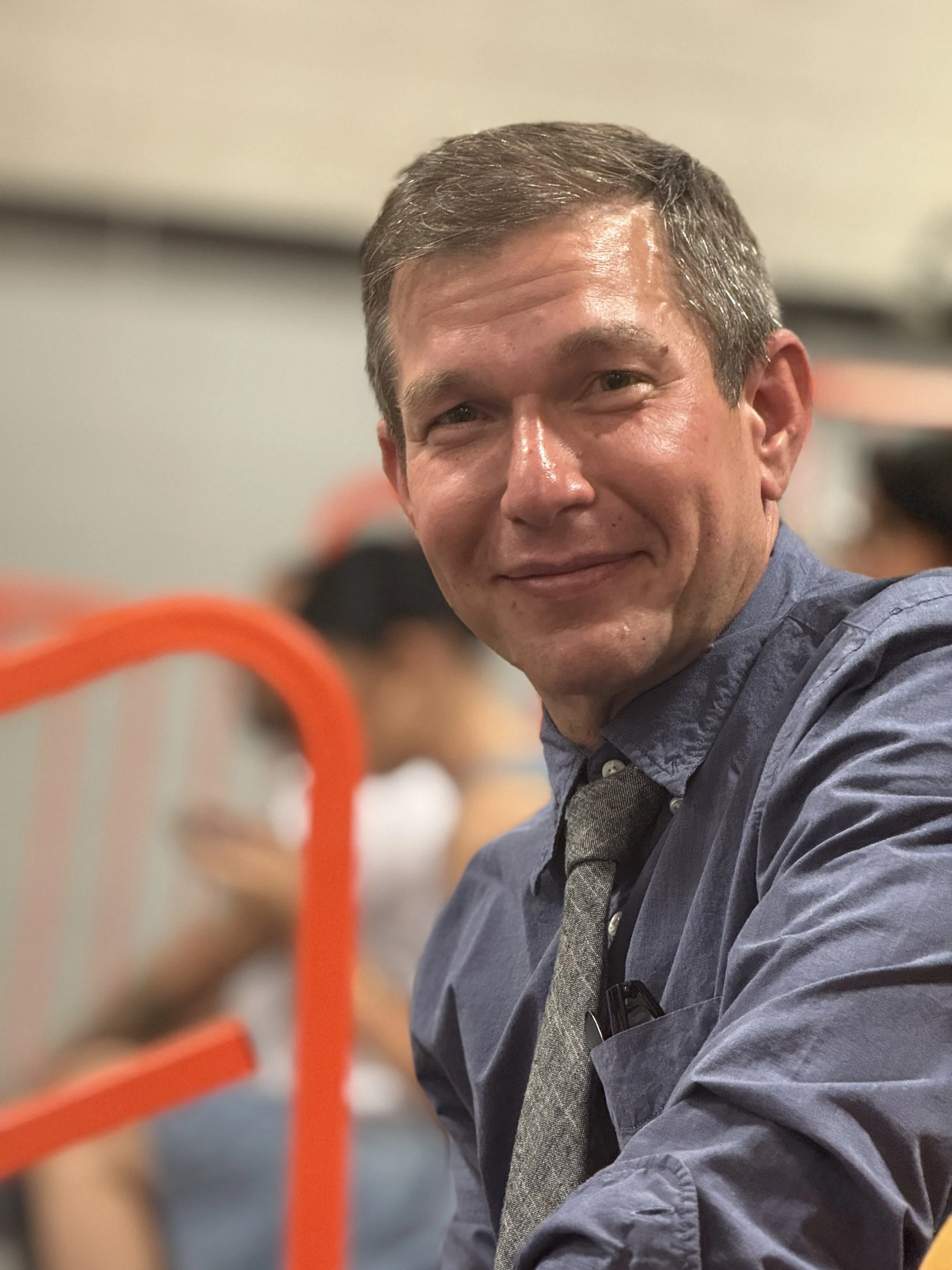 A man smiling at the camera, wearing a blue shirt and a gray tie, with people and a red railing blurred in the background.