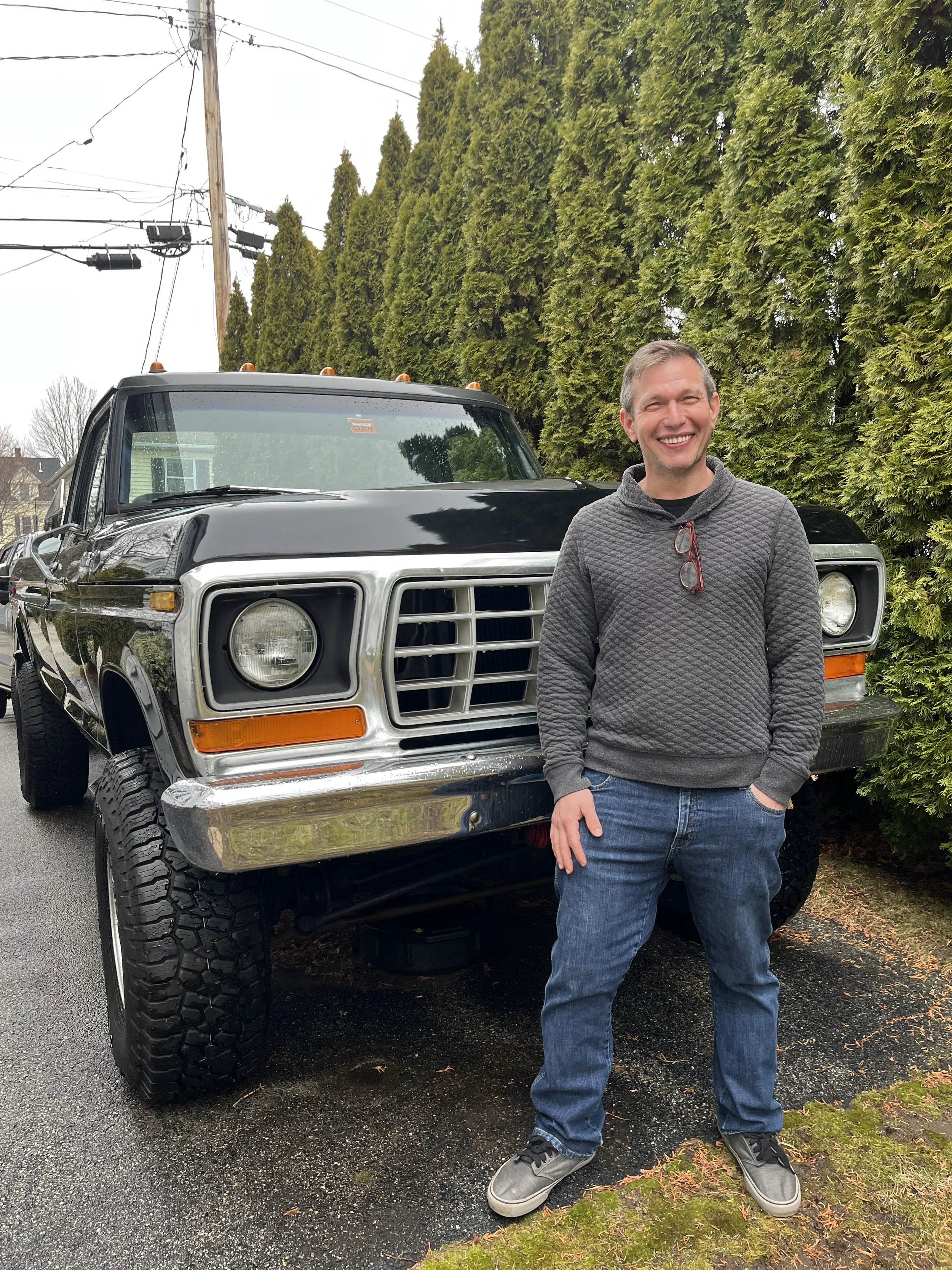 Smiling man standing in front of a large black truck with a chrome grille, on a wet driveway with green hedges and trees in the background.