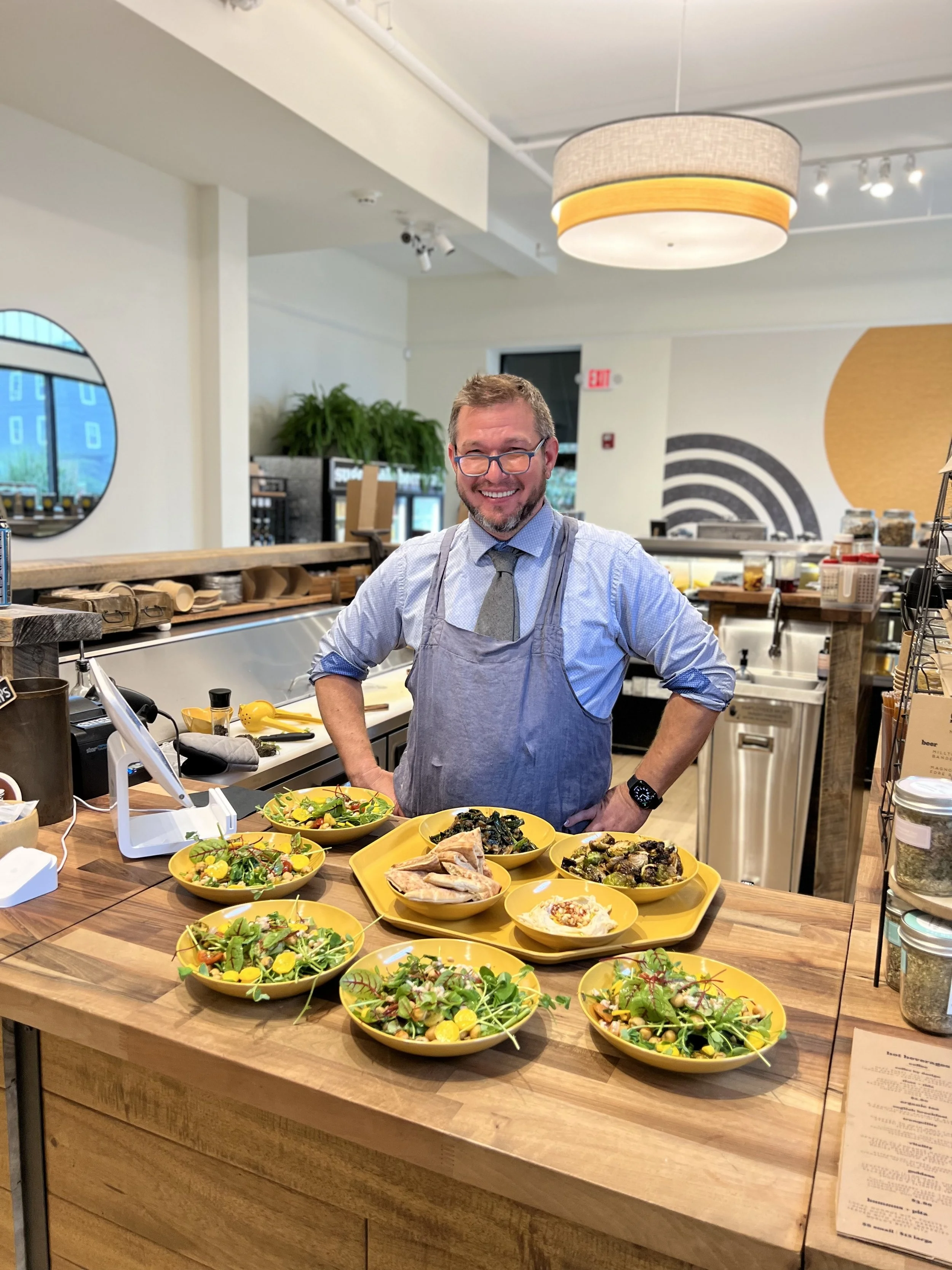 A smiling man wearing glasses, a blue shirt, and a gray apron standing behind a wooden counter with yellow bowls of salads and grilled chicken in a bright, modern cafe or restaurant.