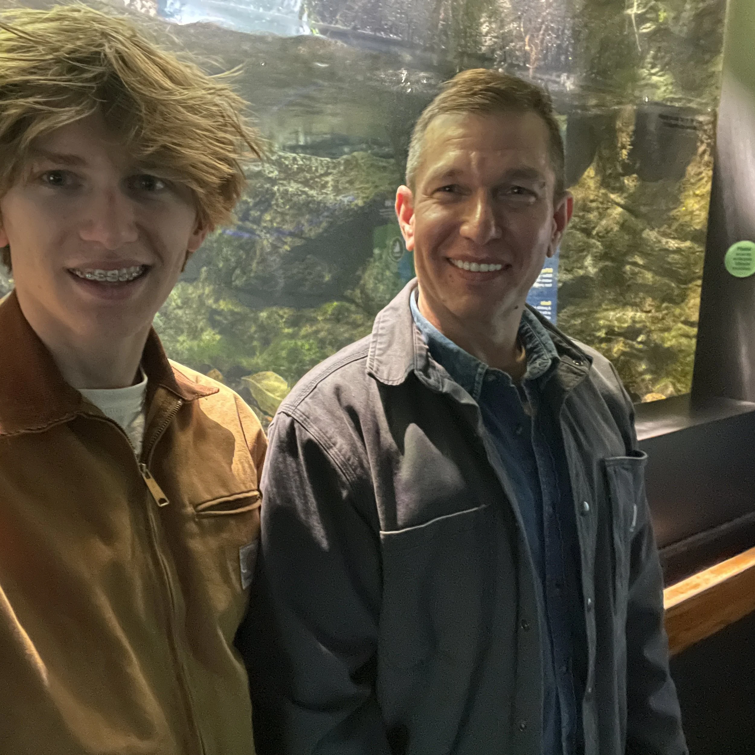 Two men standing in front of an aquarium with fish and aquatic plants visible in the background.