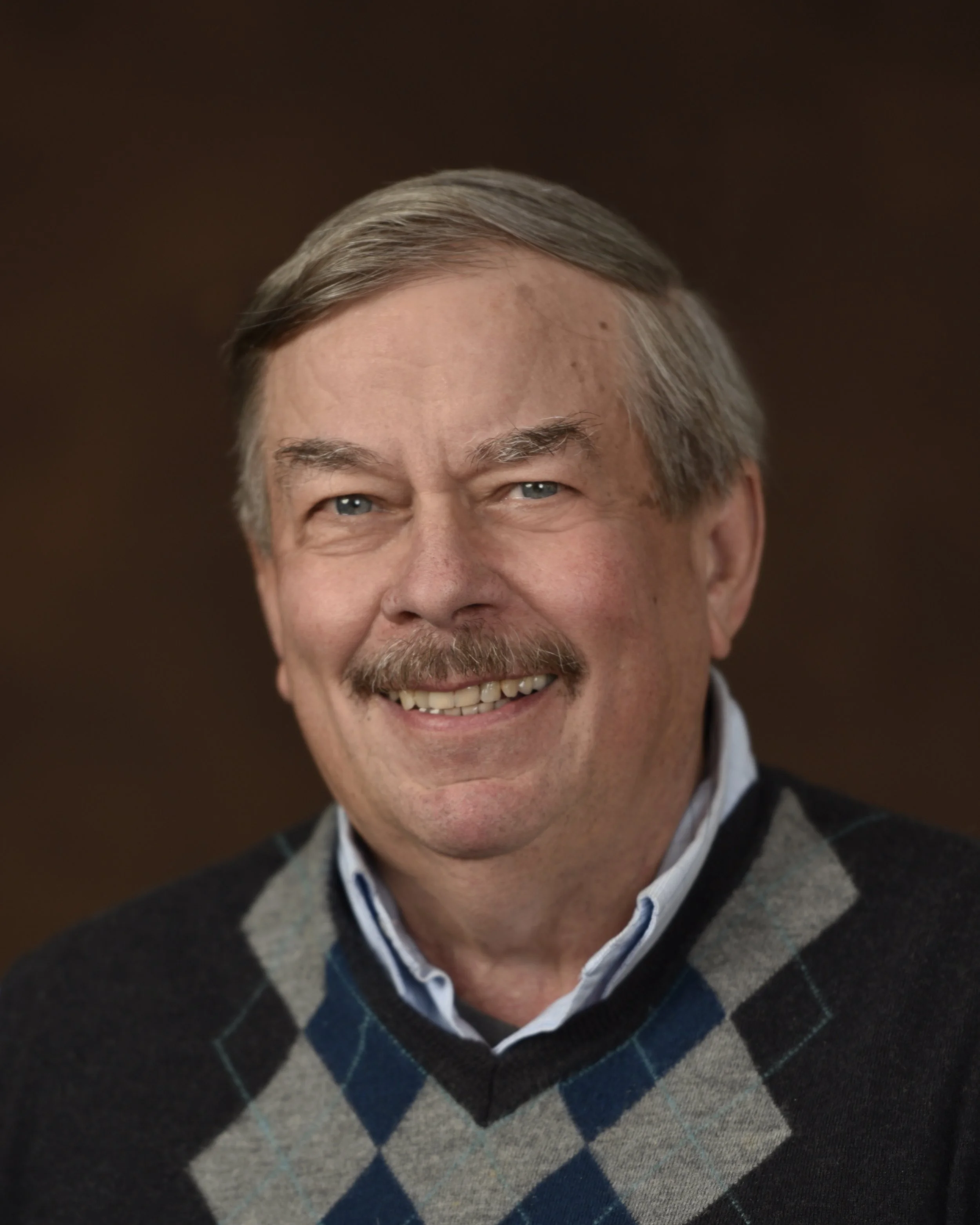 Headshot of a smiling middle-aged man with gray hair, a mustache, and blue eyes, wearing a collared shirt and argyle sweater, against a dark brown background.