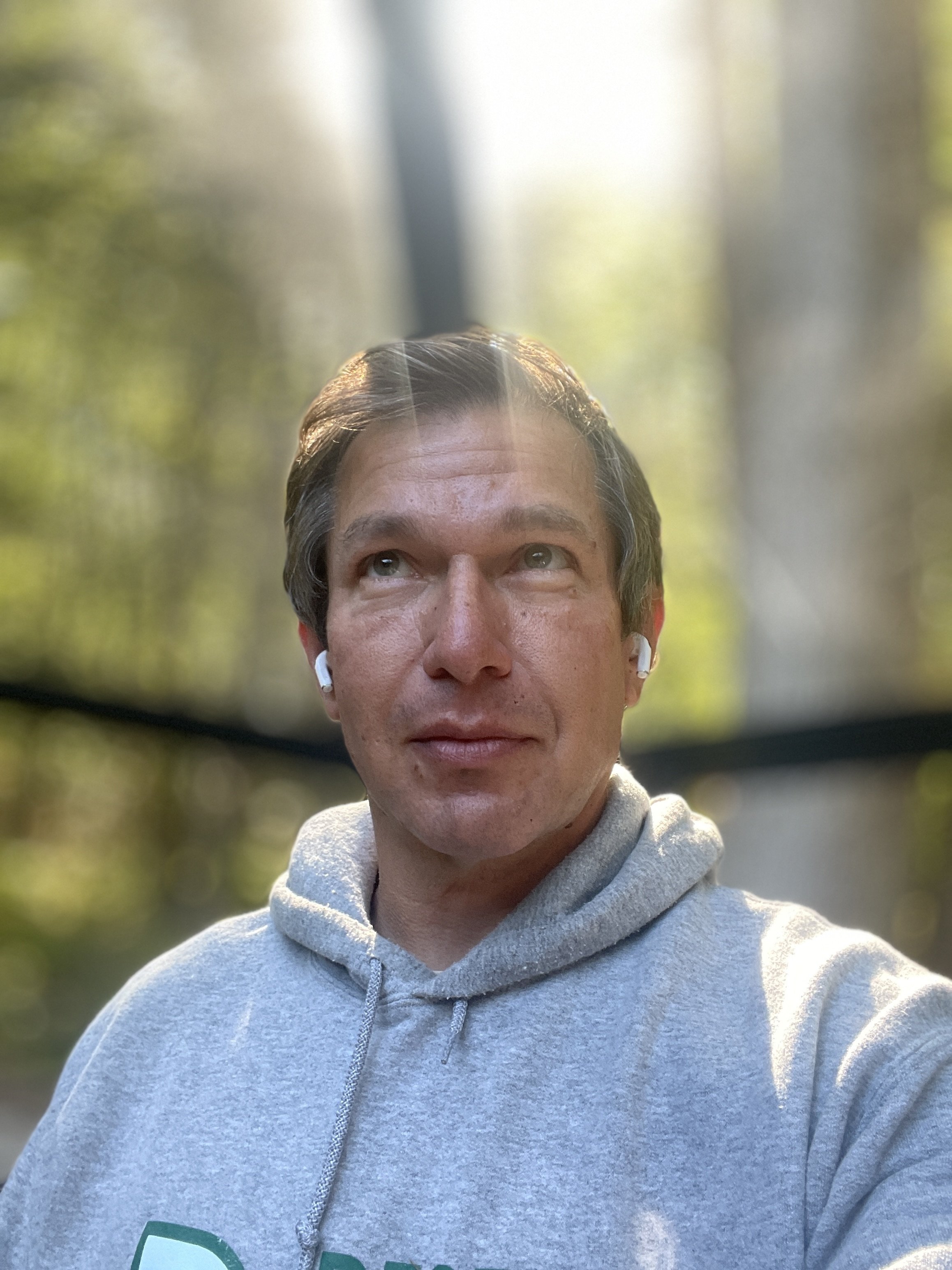 A man, named Julian Schlaver, taking a selfie outdoors with sunlight shining through trees in the background. He has dark hair, is wearing a light gray hoodie, and wireless earbuds.