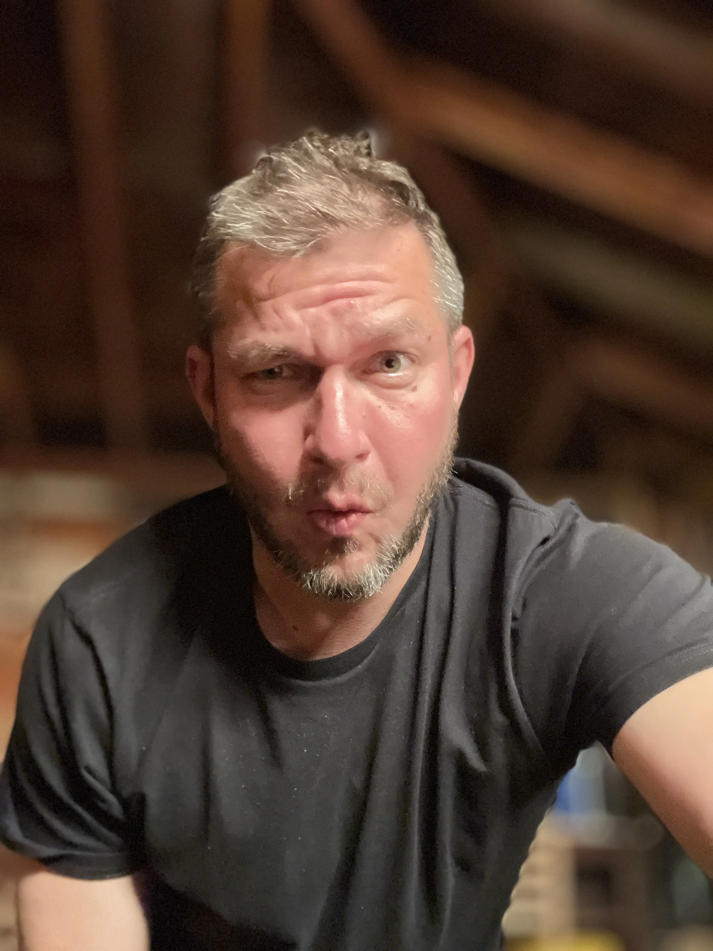 A man, named Julian Schlaver, with short gray hair and a beard making a surprised or playful face, wearing a black T-shirt, in a dimly lit indoor setting with wooden beams.