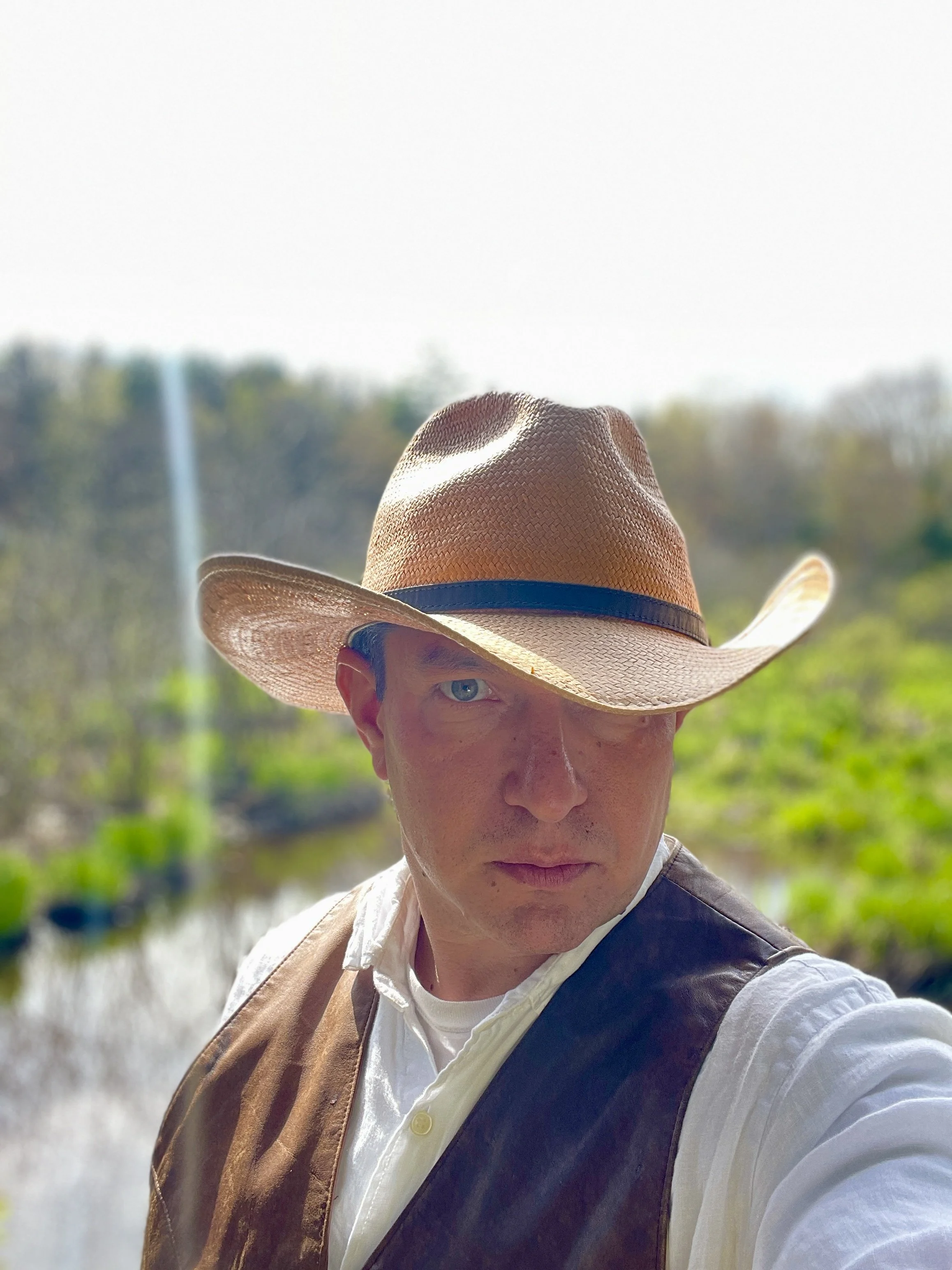 A man, named Julian Schlaver, in a white shirt, brown vest, and wide-brimmed hat standing outdoors near a body of water with a waterfall in the background.