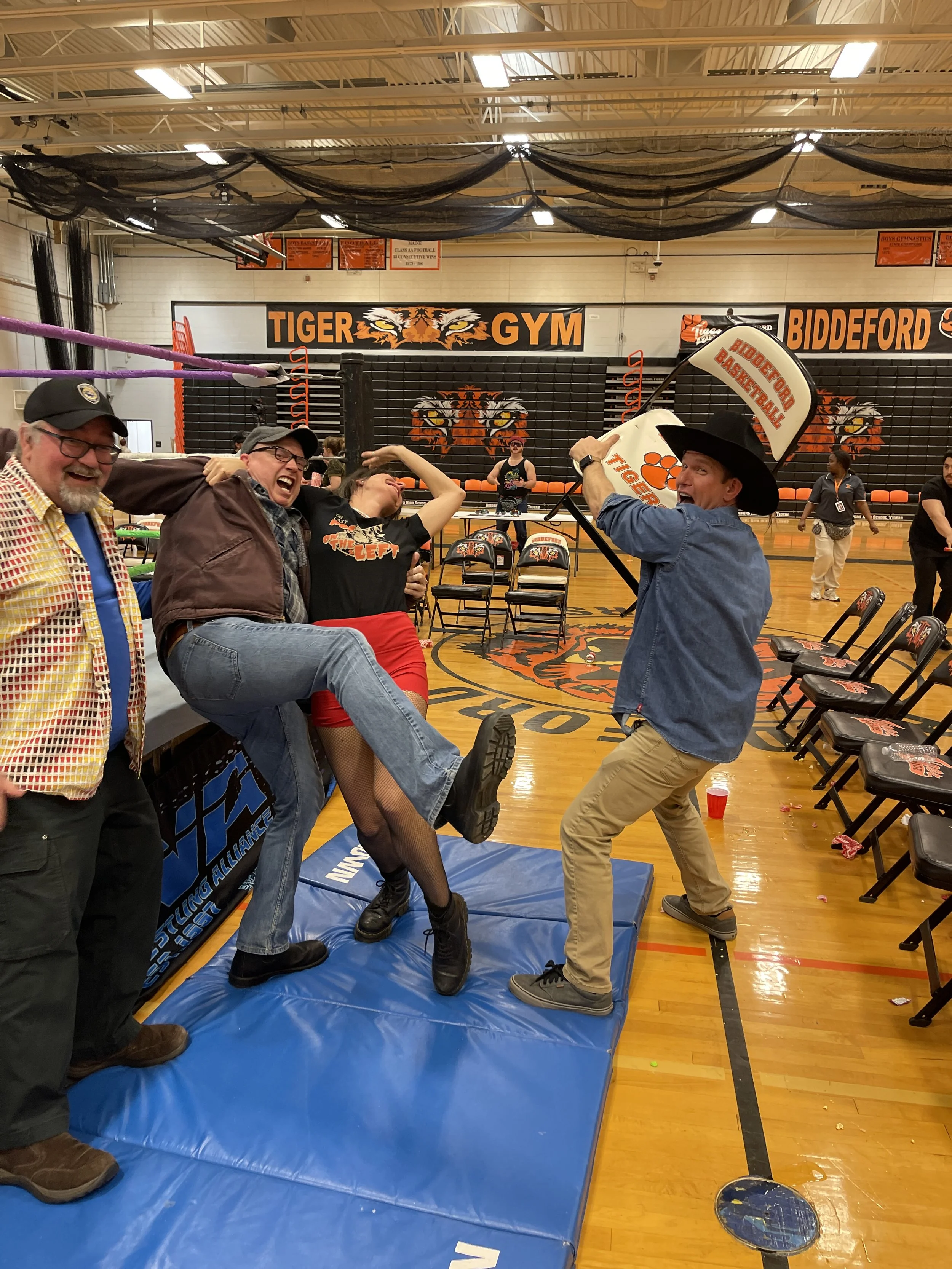 People having fun and playfully pretending to fight with foam bats in a school gym decorated in black and orange tiger theme, with chairs set up and a banner that reads 'Tiger Gym' and 'Biddeford' in the background.
