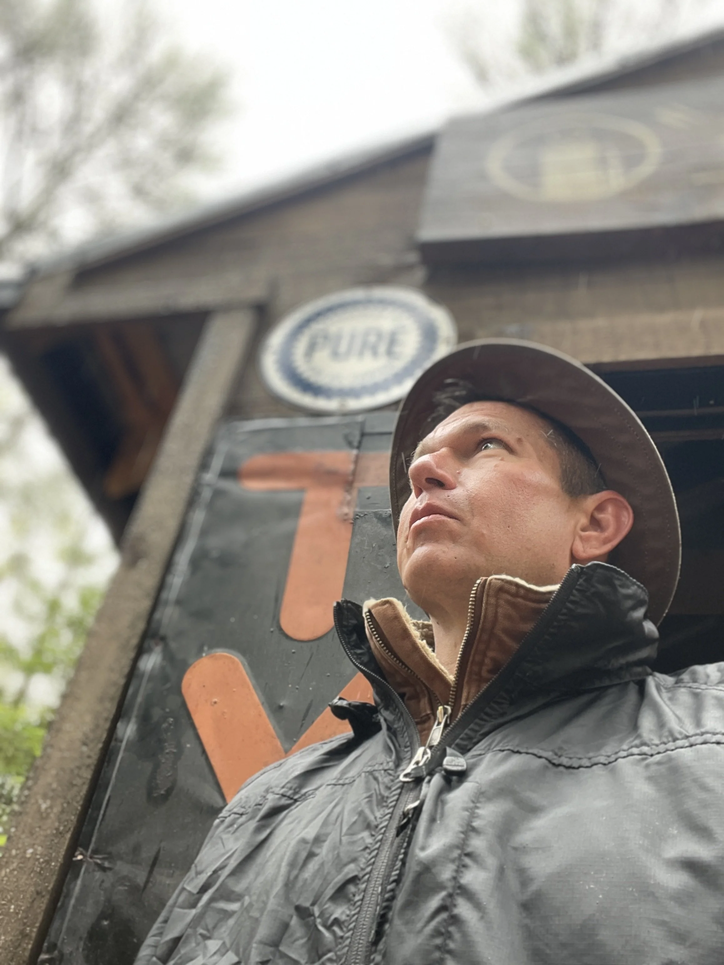 Man wearing a brown hat and black jacket looking up outside near a building with a «PURE» sign