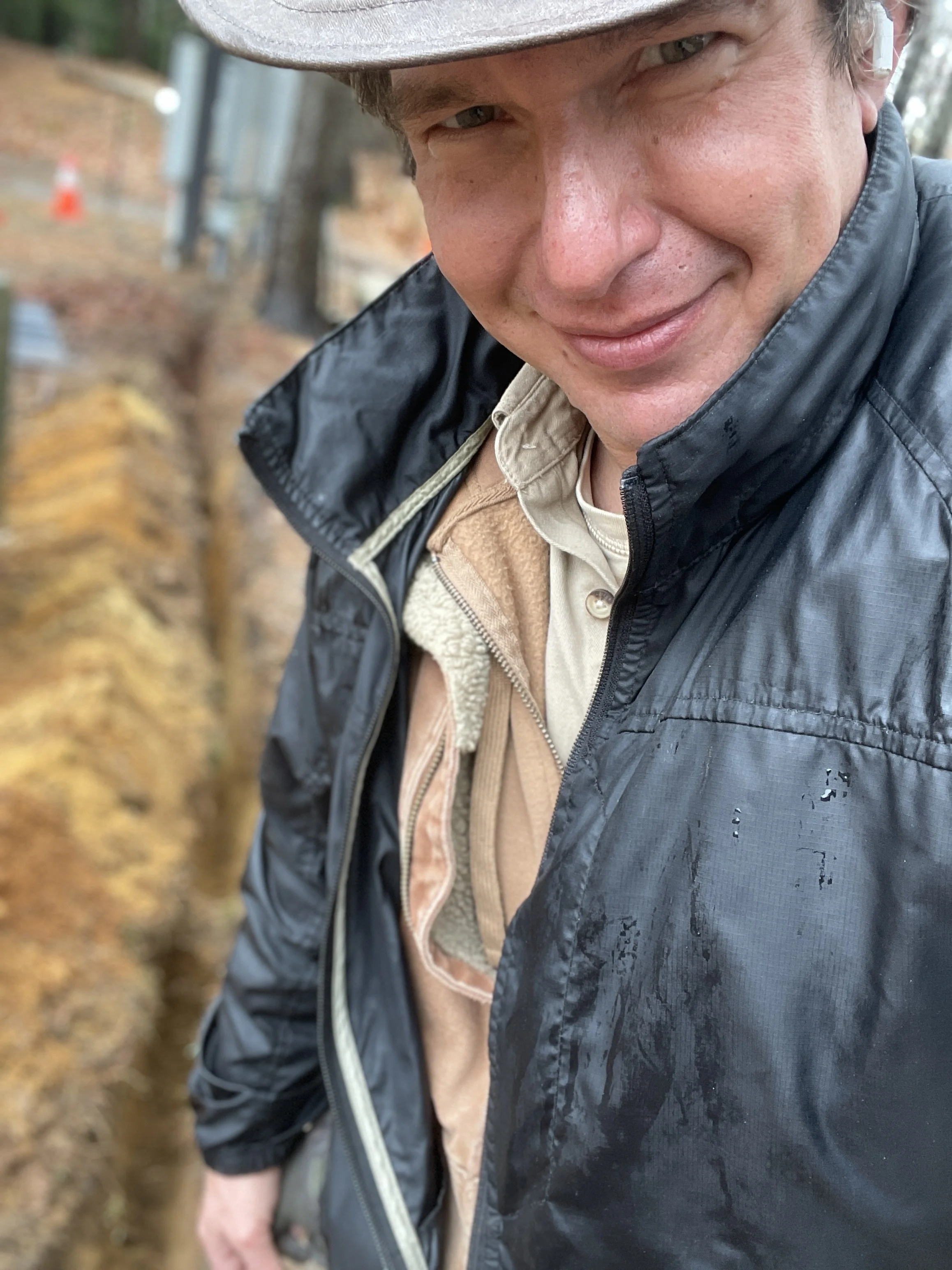 Close-up of a man, named Julian Schlaver, outdoors wearing a tan hat, layered clothing, and a black waterproof jacket, smiling at the camera with autumn leaves in the background.