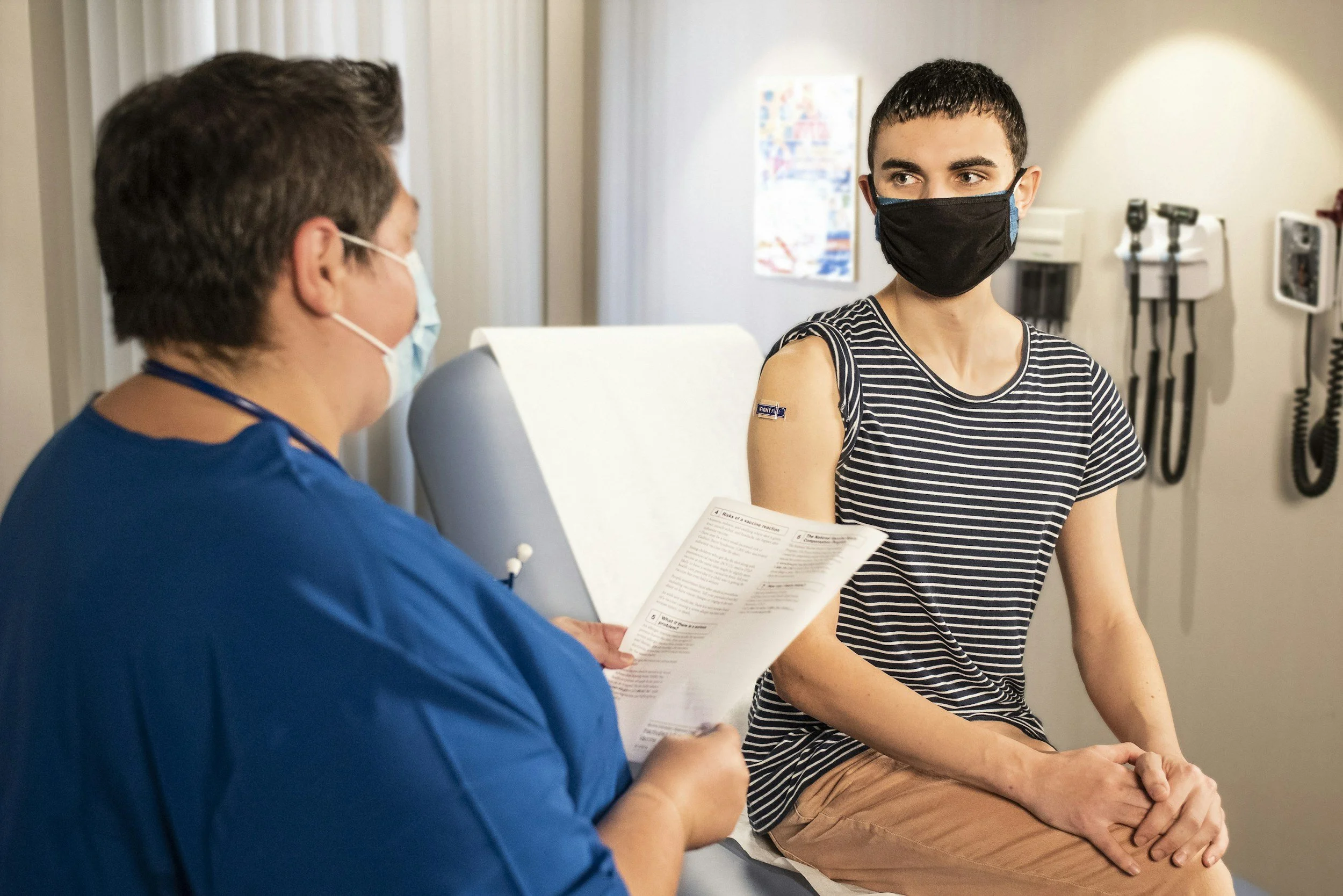 A young man sitting in a medical examination room, receiving a vaccination from a healthcare professional. Both are wearing face masks, and the young man is dressed in a striped shirt with his sleeve rolled up.