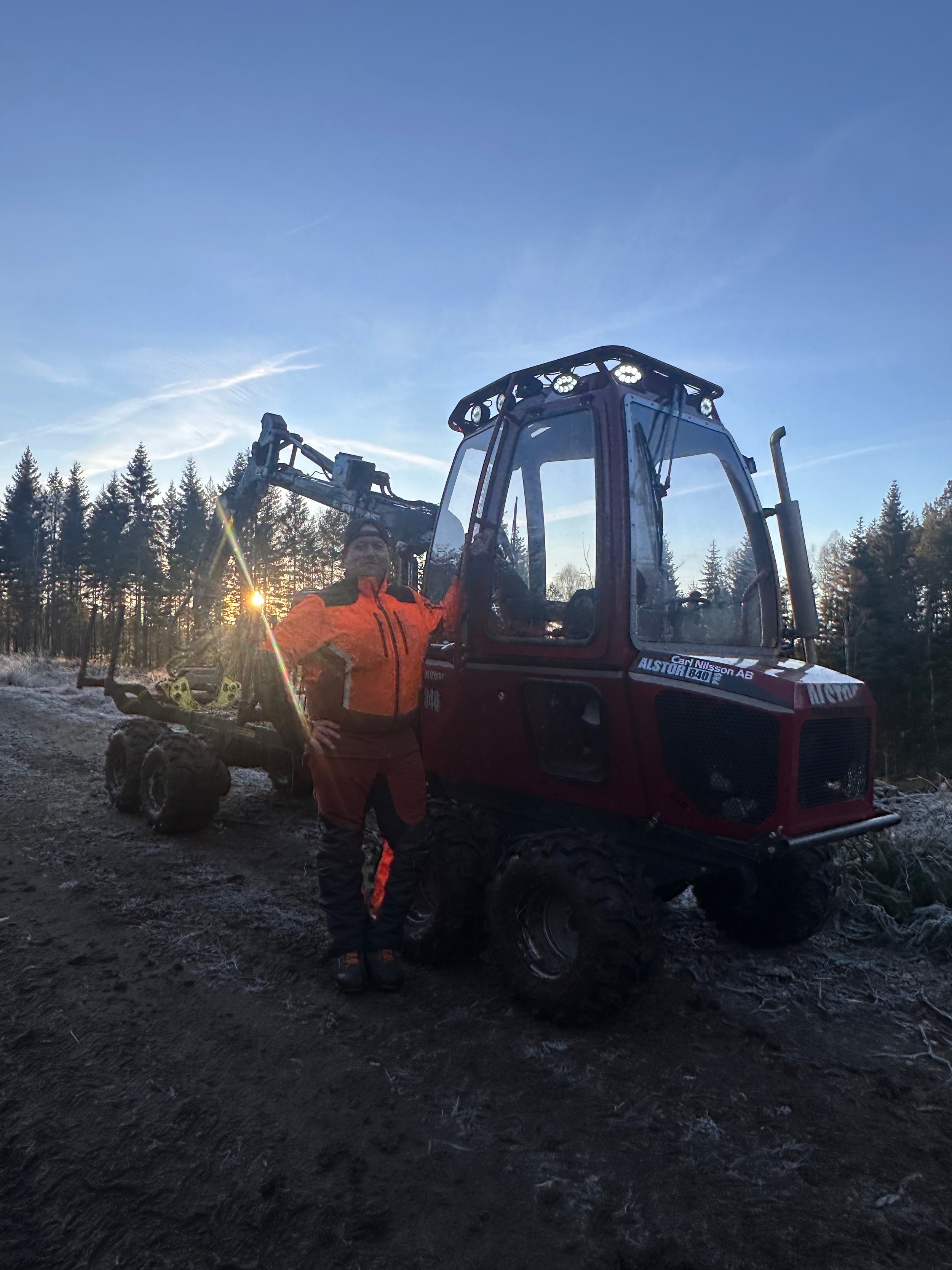 Person i orange och svarta skyddskläder står bredvid ett rött skogsbruksfordon på en grusväg med träd i bakgrunden och solnedgången.