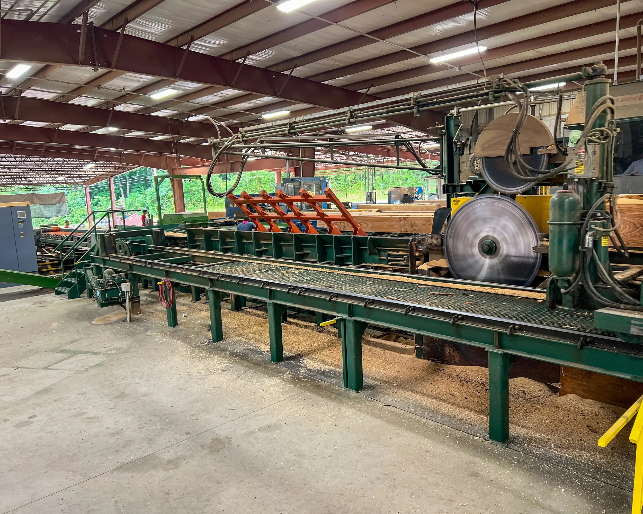 Wood processing machine in a lumber mill, with saw blades cutting wood and conveyor belt transporting lumber inside a large industrial building.