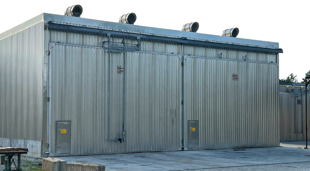 A large industrial metal building with metal siding and a sliding door, equipped with vents on top and electrical panels at the bottom.