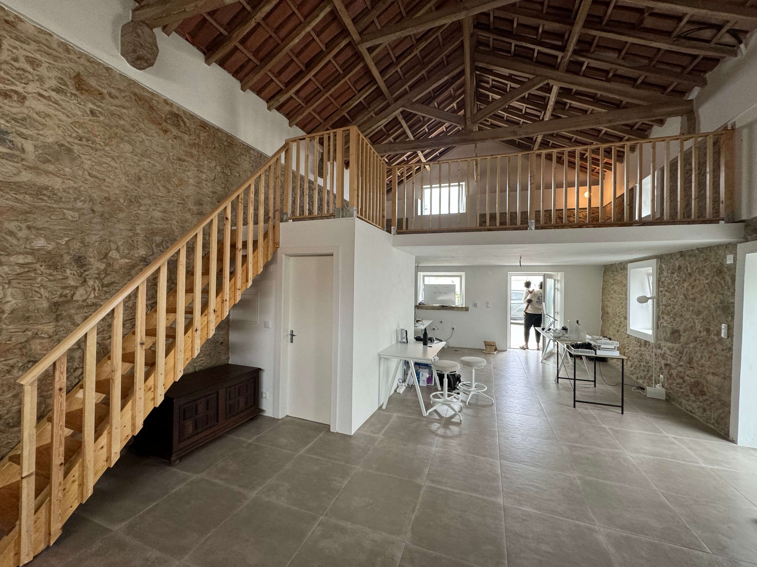 Interior of a spacious room with stone walls, tiled floor, wooden ceiling, and a mezzanine level with wooden railing. There is a staircase on the left leading to the upper level, white furniture, and a person standing near the door at the back.