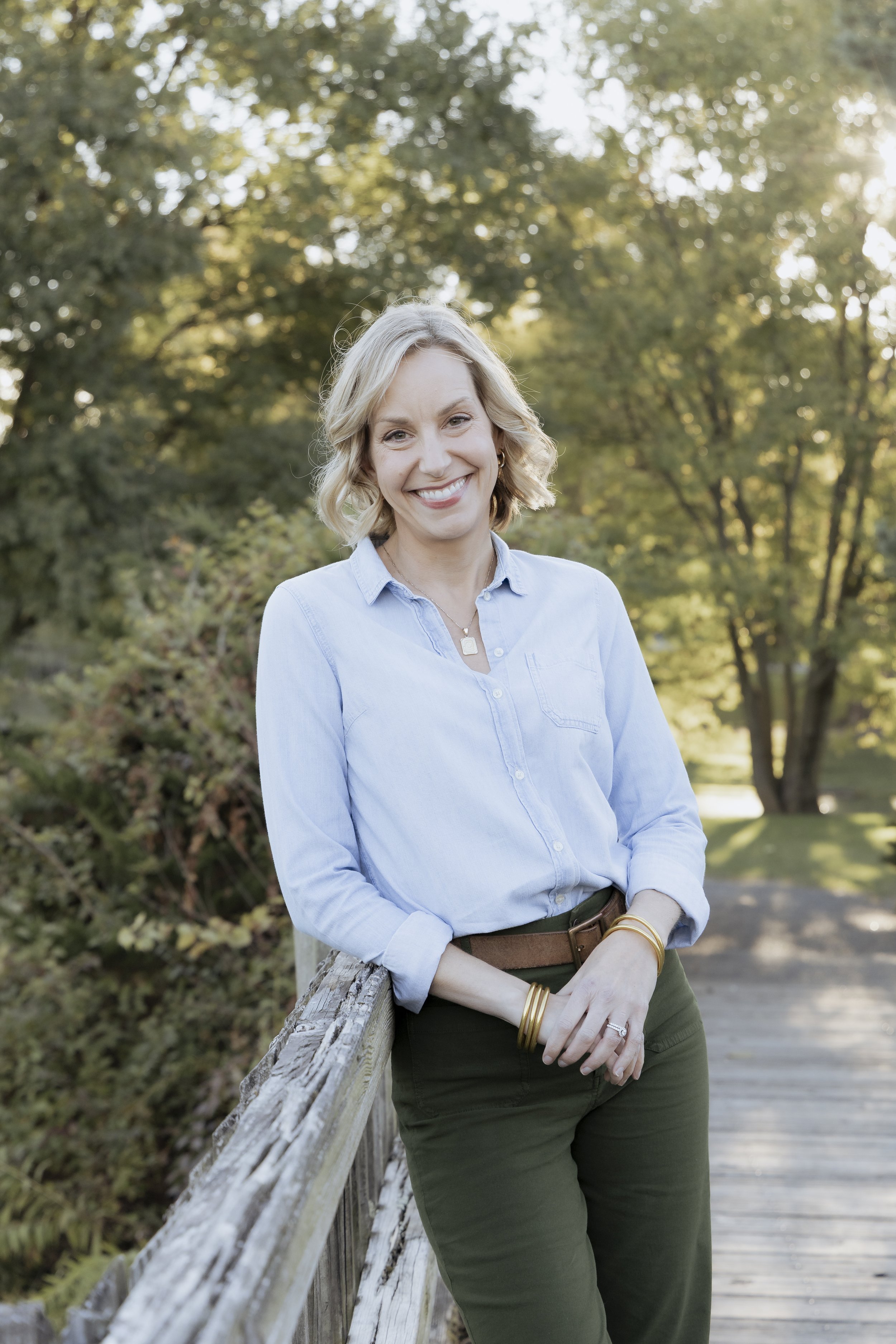 A smiling woman with blonde hair in a bob, wearing a light blue button-up shirt, dark green pants with a brown belt, and gold bracelets, leaning on a wooden railing in a park with green trees and sunlight in the background.