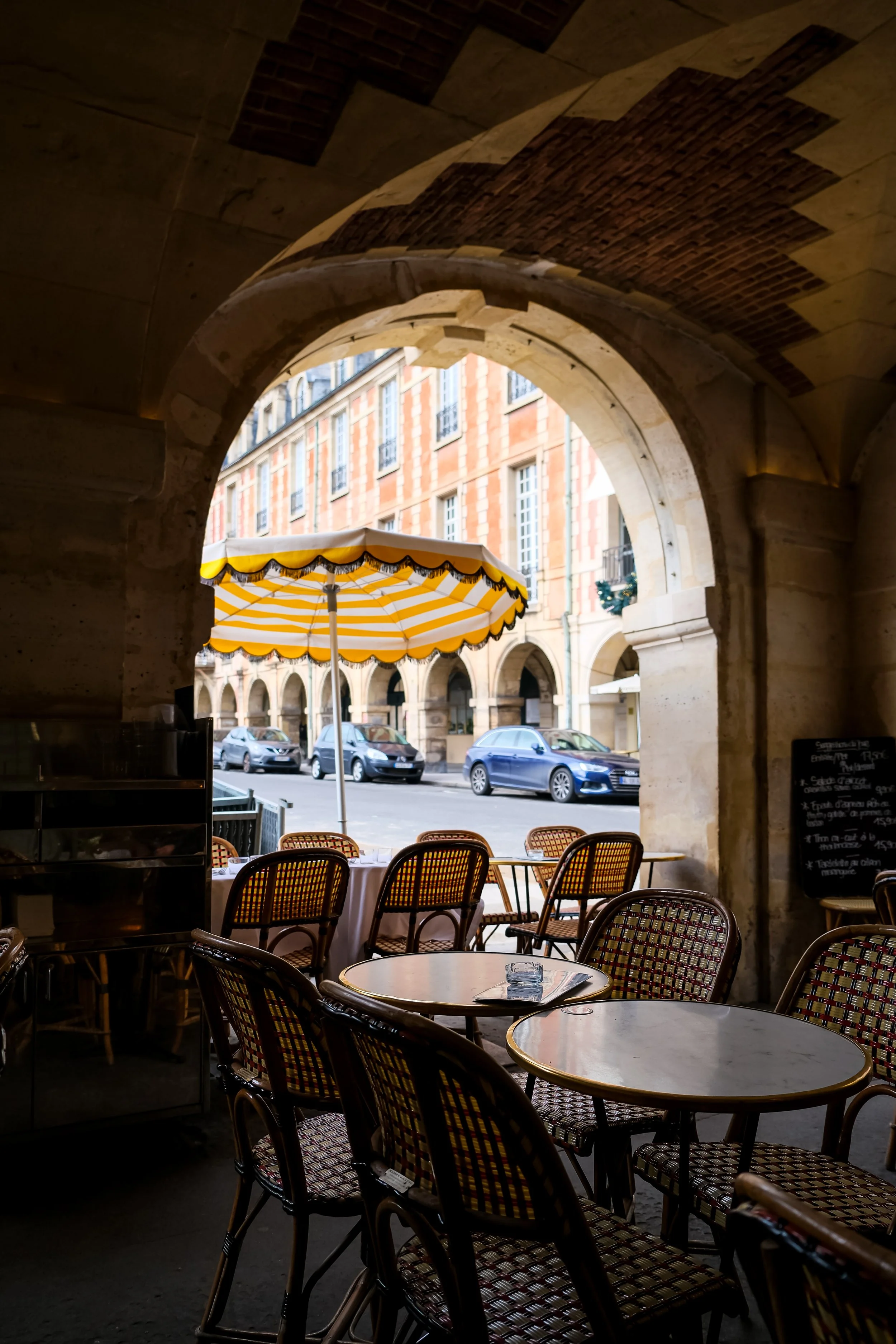 Outdoor cafe with yellow and white striped umbrella, wooden chairs with woven seats, and round tables, framed by a stone archway, street with parked cars, and historic buildings in the background.