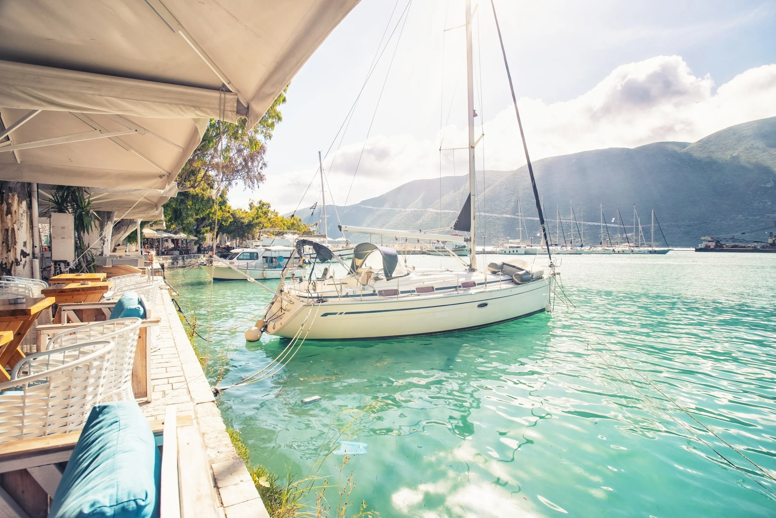 A marina with boats docked near a waterfront restaurant, with a mountain range in the background and umbrellas shading outdoor seating.