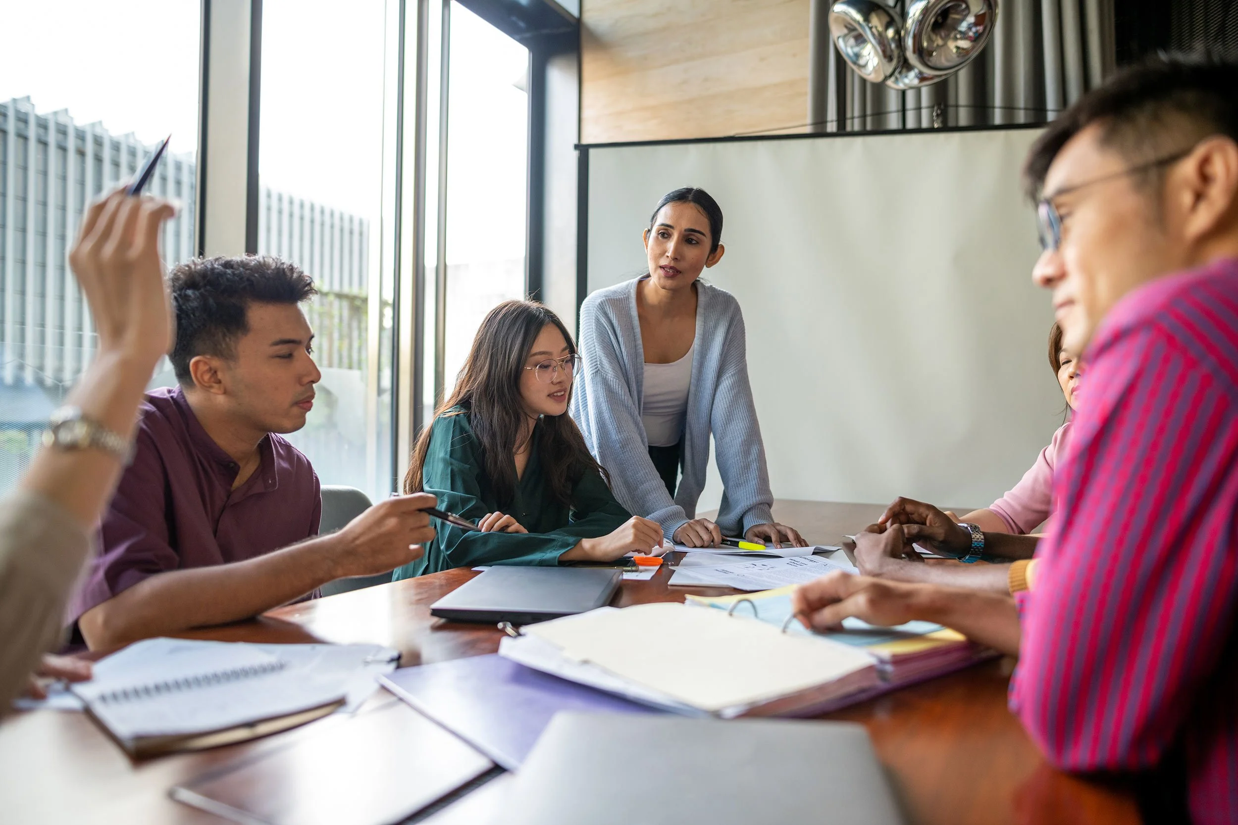 A person standing at a conference table leading a group discussion with four colleagues reviewing documents