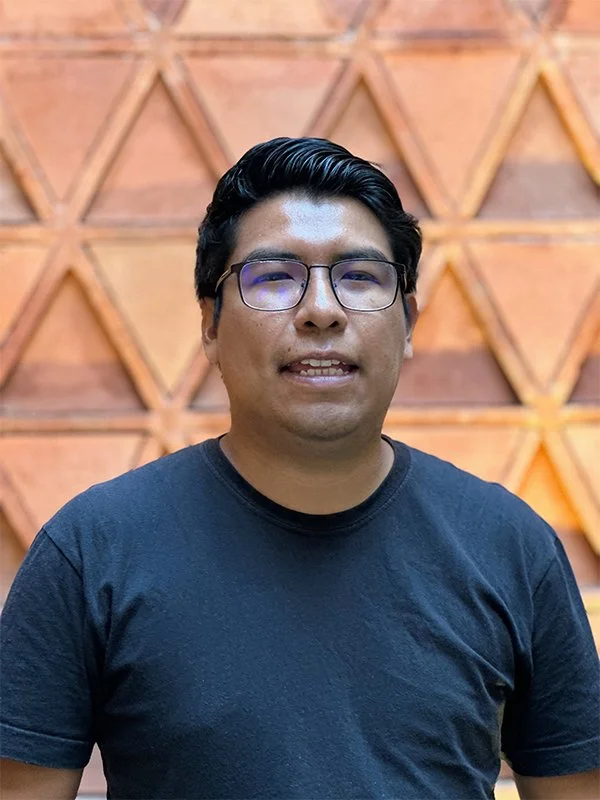 Joven hombre con gafas y camiseta negra posando frente a una pared de madera con patrón triangular