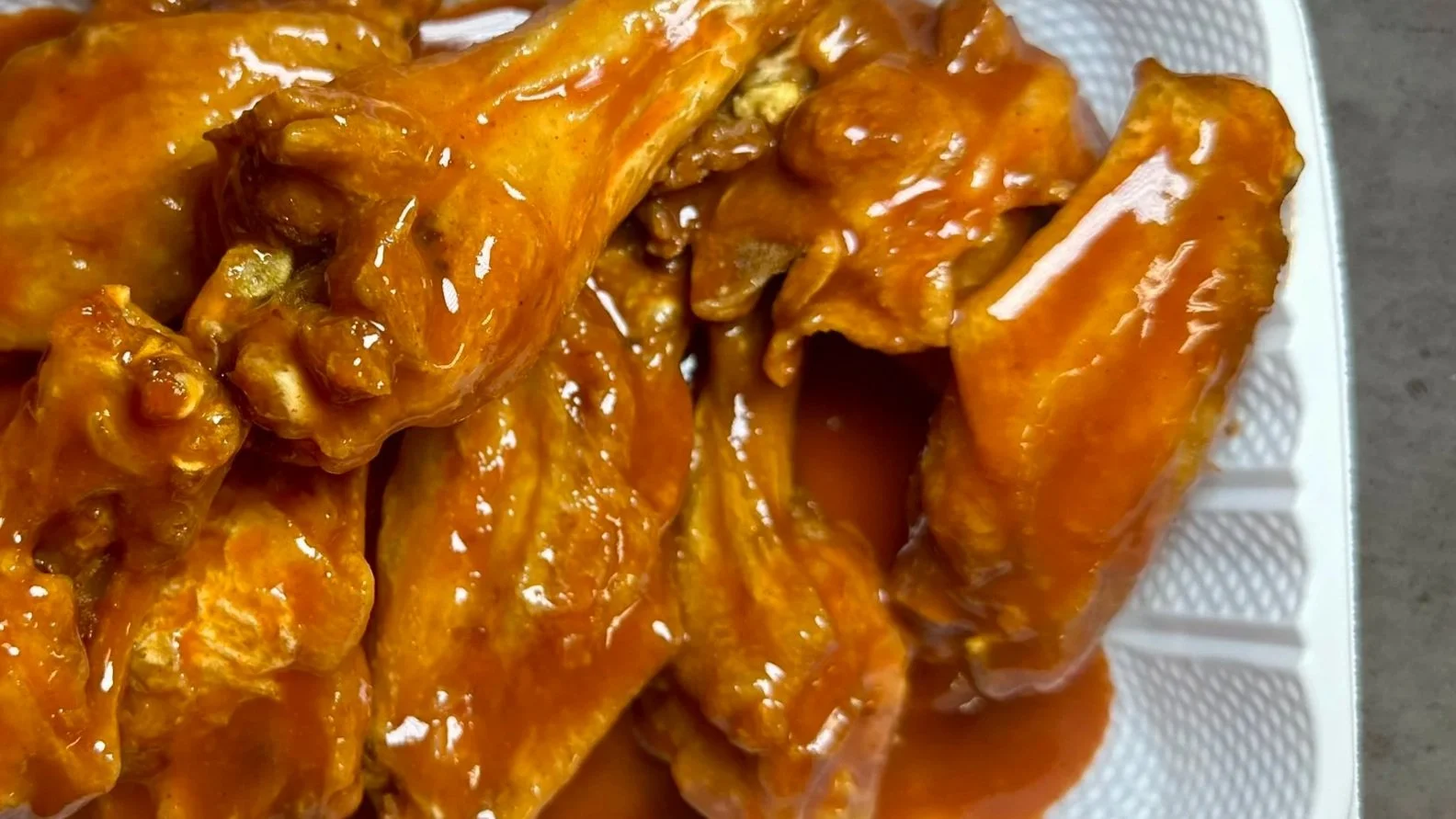 Six orange glazed chicken wings inside a white takeout container on a gray surface.