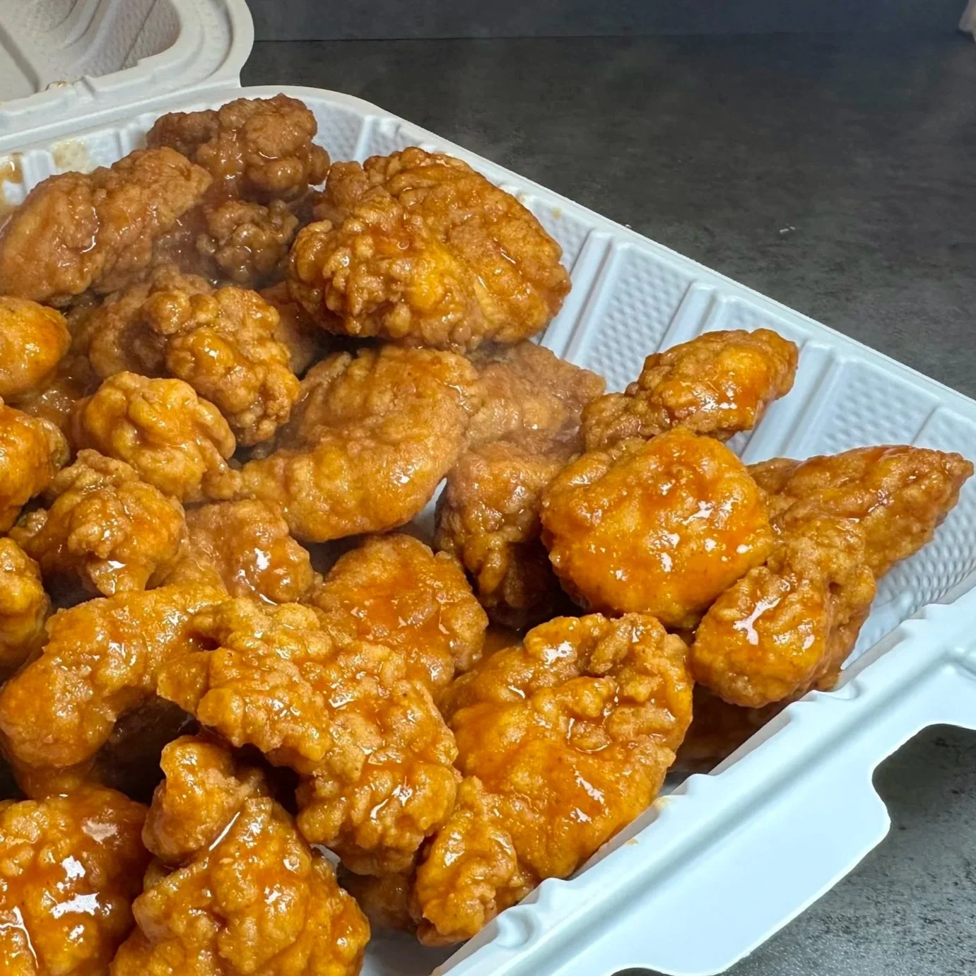 Takeout container filled with orange chicken pieces covered in sauce, placed on a dark countertop.