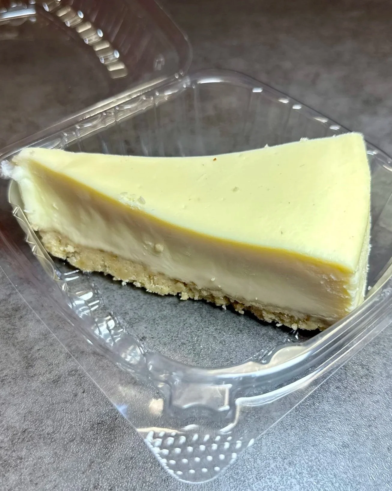 A slice of cheesecake with a graham cracker crust in a clear plastic container on a gray surface.