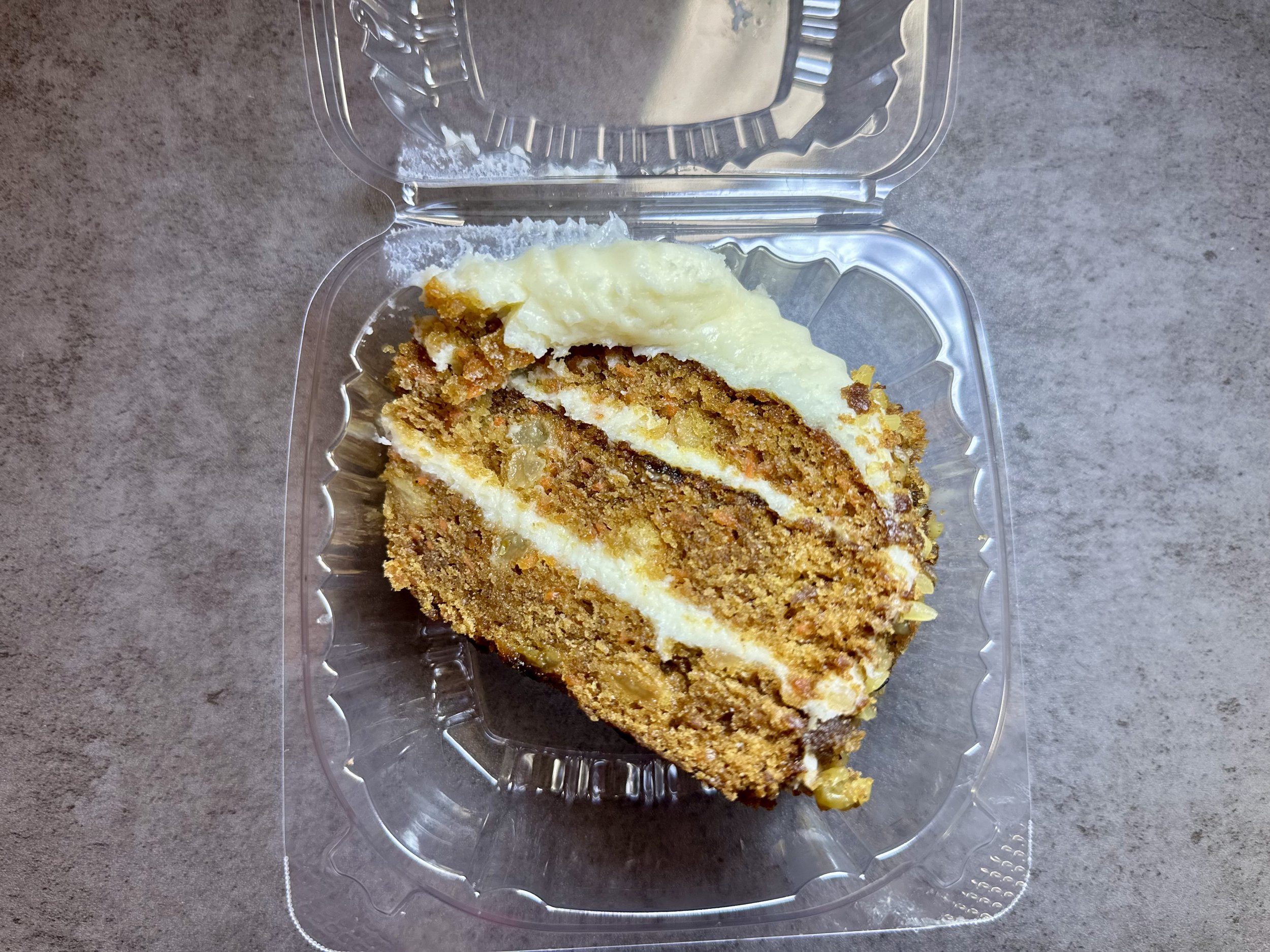 Slice of carrot cake with cream cheese frosting in a clear plastic container on a gray surface.