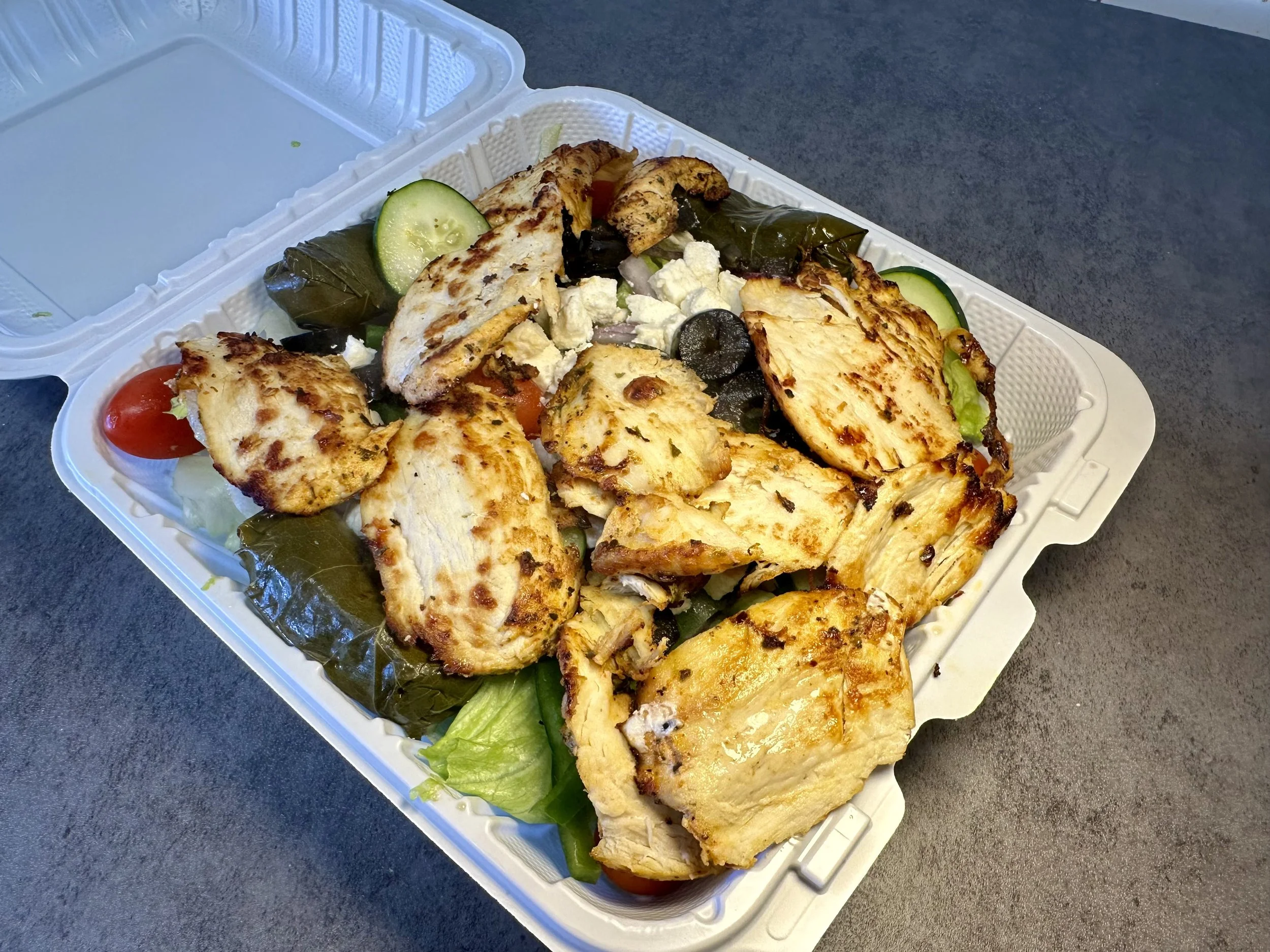 Grilled chicken salad with cucumbers, cherry tomatoes, black olives, feta cheese, and lettuce in a takeout container.