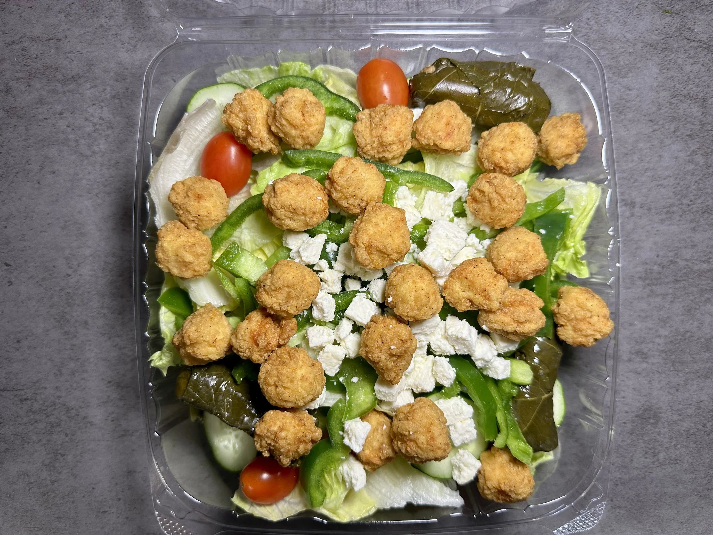 Container of salad with fried chicken, cherry tomatoes, cucumbers, green bell peppers, lettuce, pickles, and crumbled cheese.