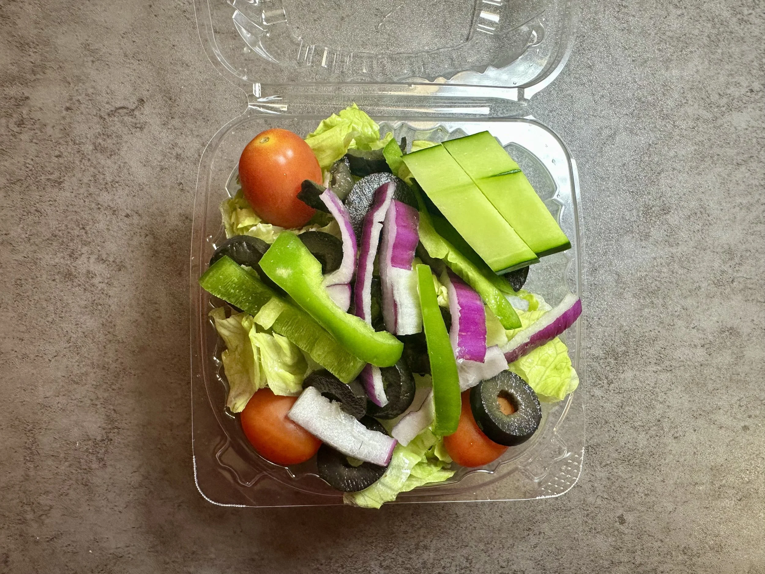 Plastic container of salad with cherry tomatoes, green bell peppers, red onions, sliced cucumbers, sliced black olives, and lettuce on a gray surface.