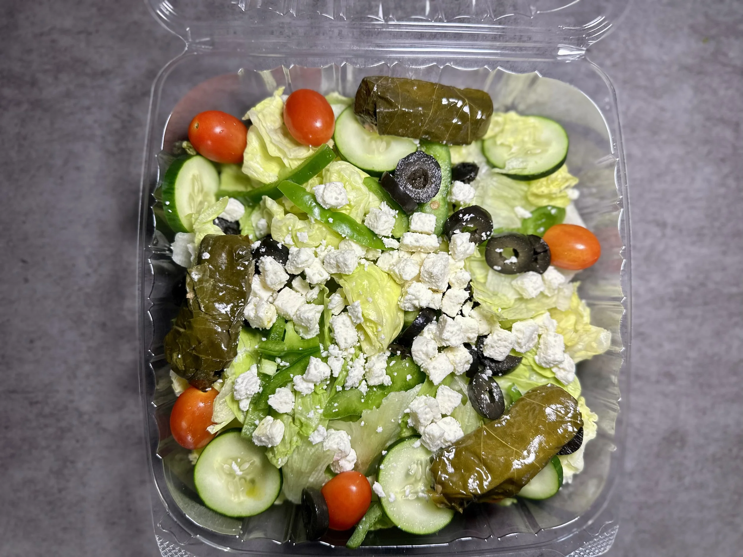 Mixed salad in a plastic container with cherry tomatoes, cucumbers, black olives, lettuce, grape leaves, crumbled feta cheese, and green peppers.