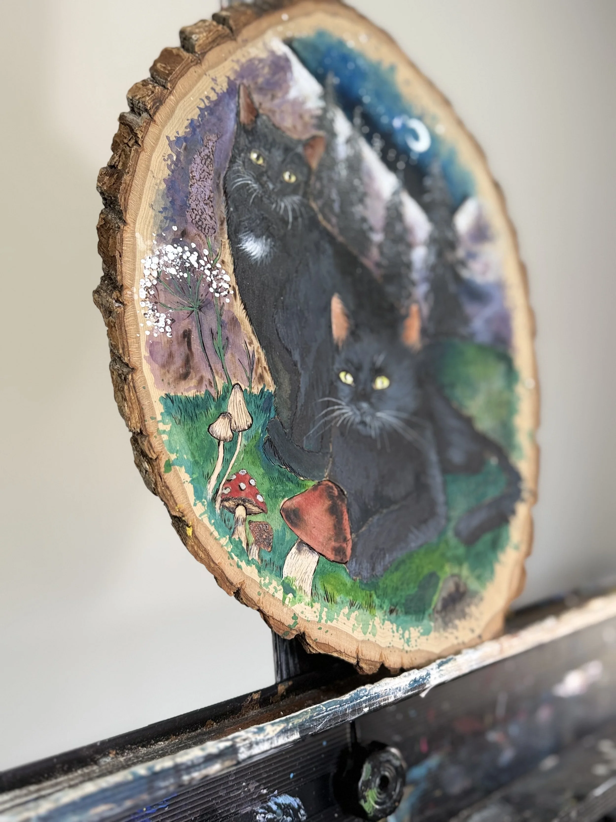 Painted wood slice featuring two black cats surrounded by colorful foliage and mushrooms.