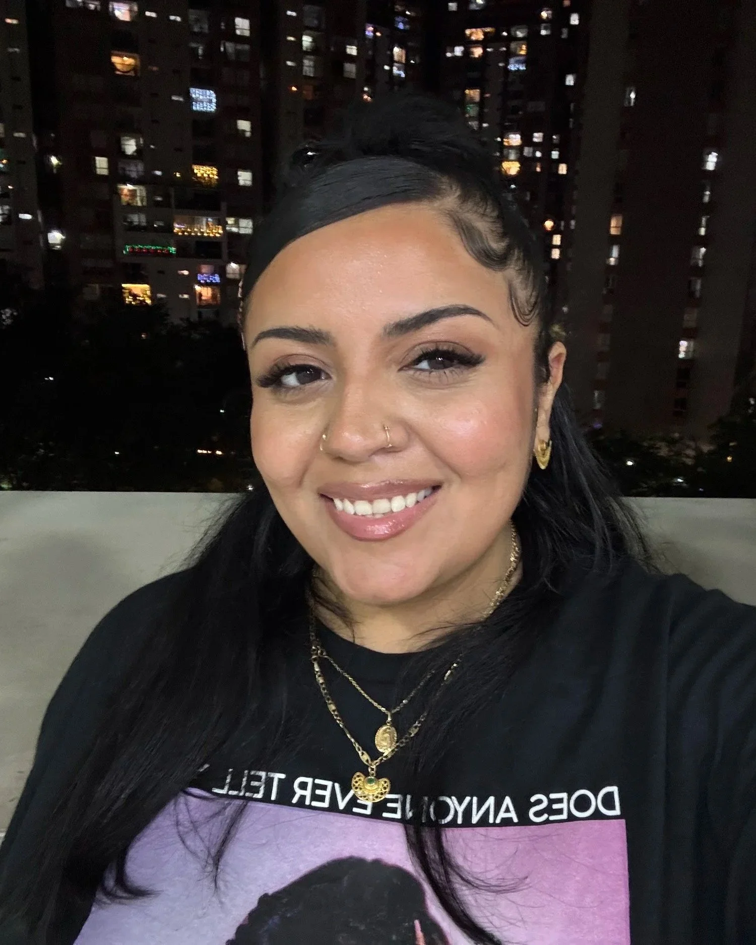 A smiling woman with black hair styled with a side parting, wearing gold jewelry, including earrings and layered necklaces. She is wearing a black message t-shirt. The background shows a cityscape at night with high-rise buildings and illuminated windows.