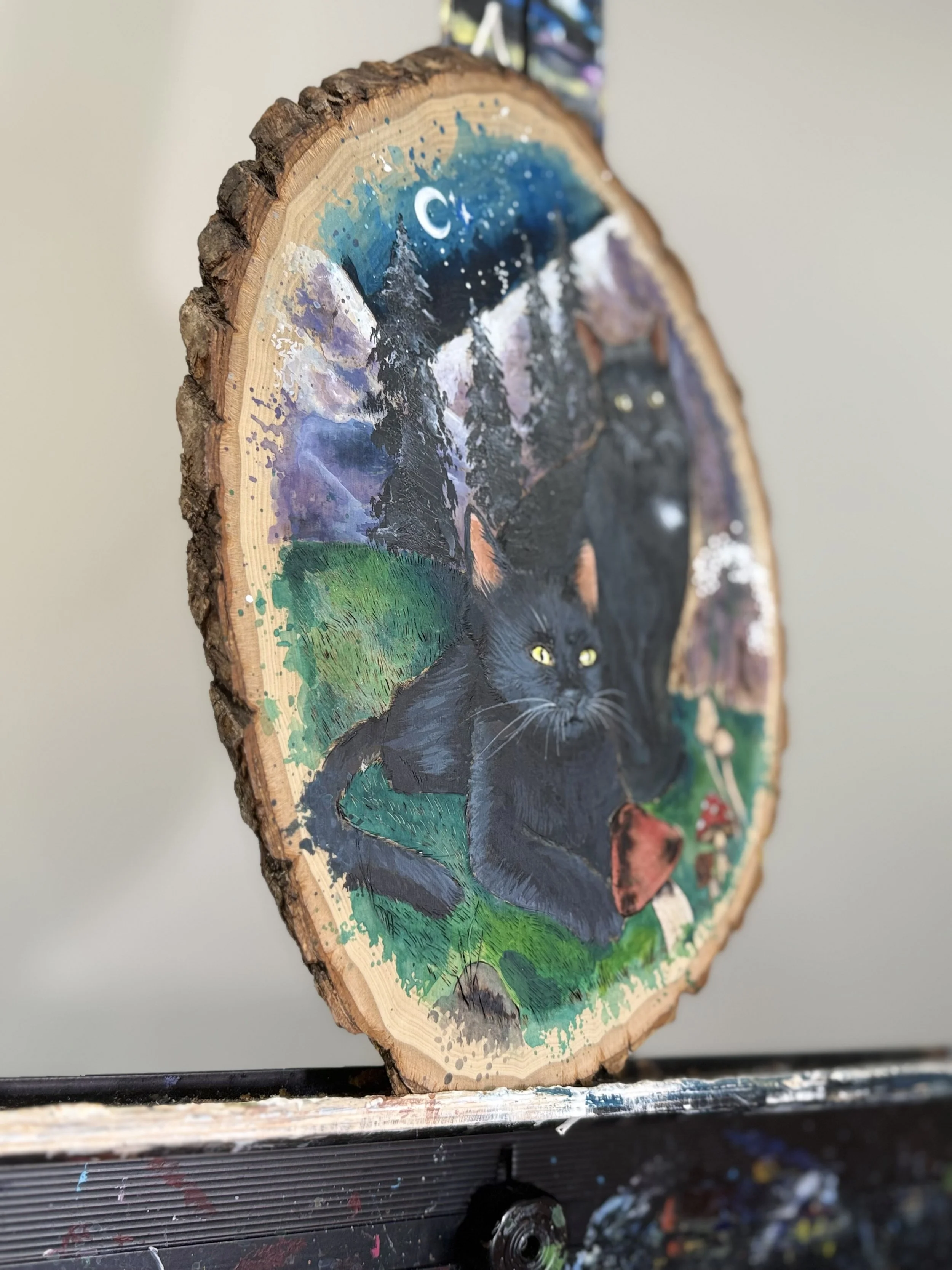 A painted wooden slice featuring a black cat in a forest with mountains, trees, a crescent moon, and a starry sky.