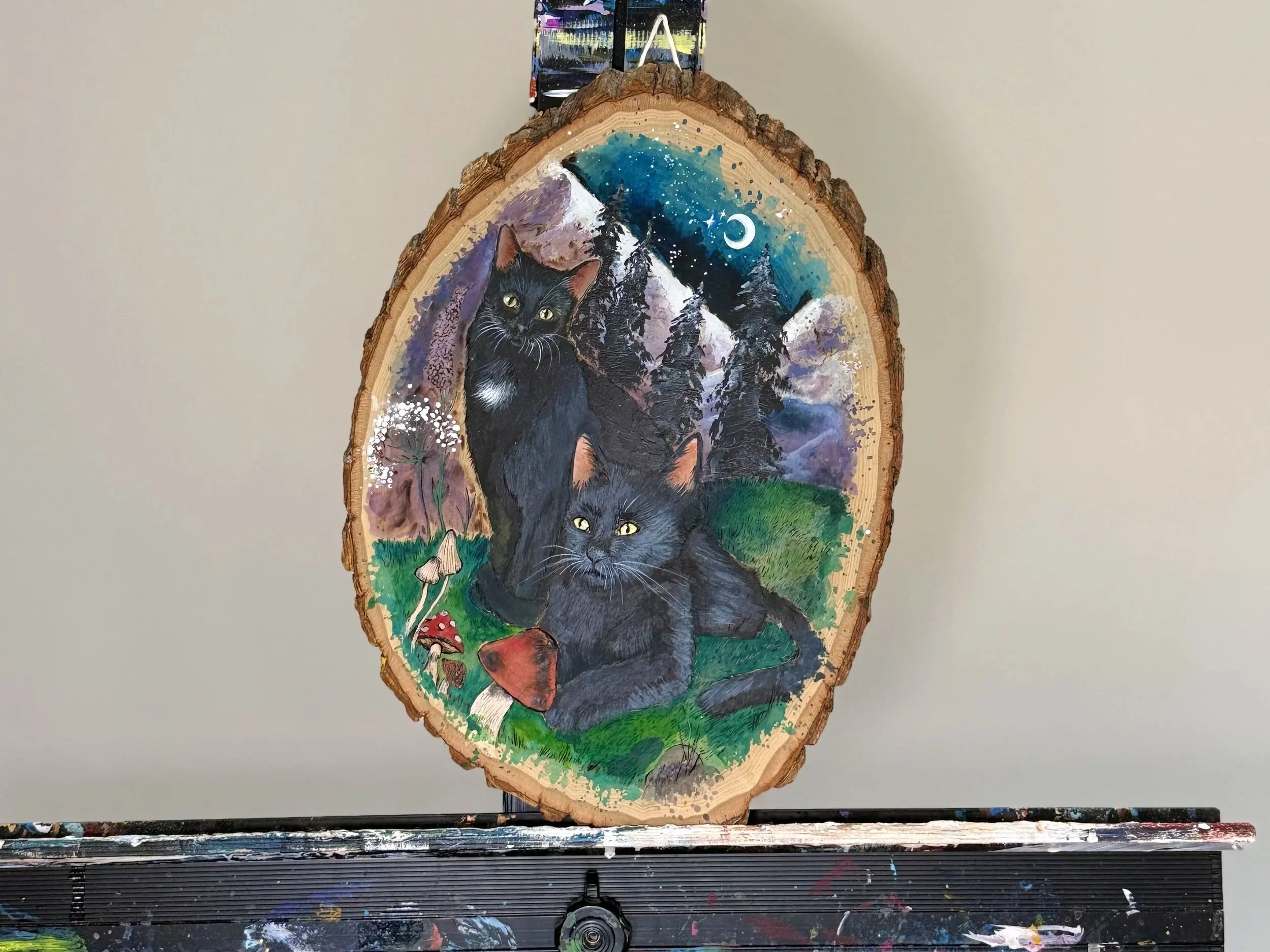 Painting of two black cats in a forest scene on a wood slice, with mountains, trees, a night sky, stars, a moon, and mushrooms.