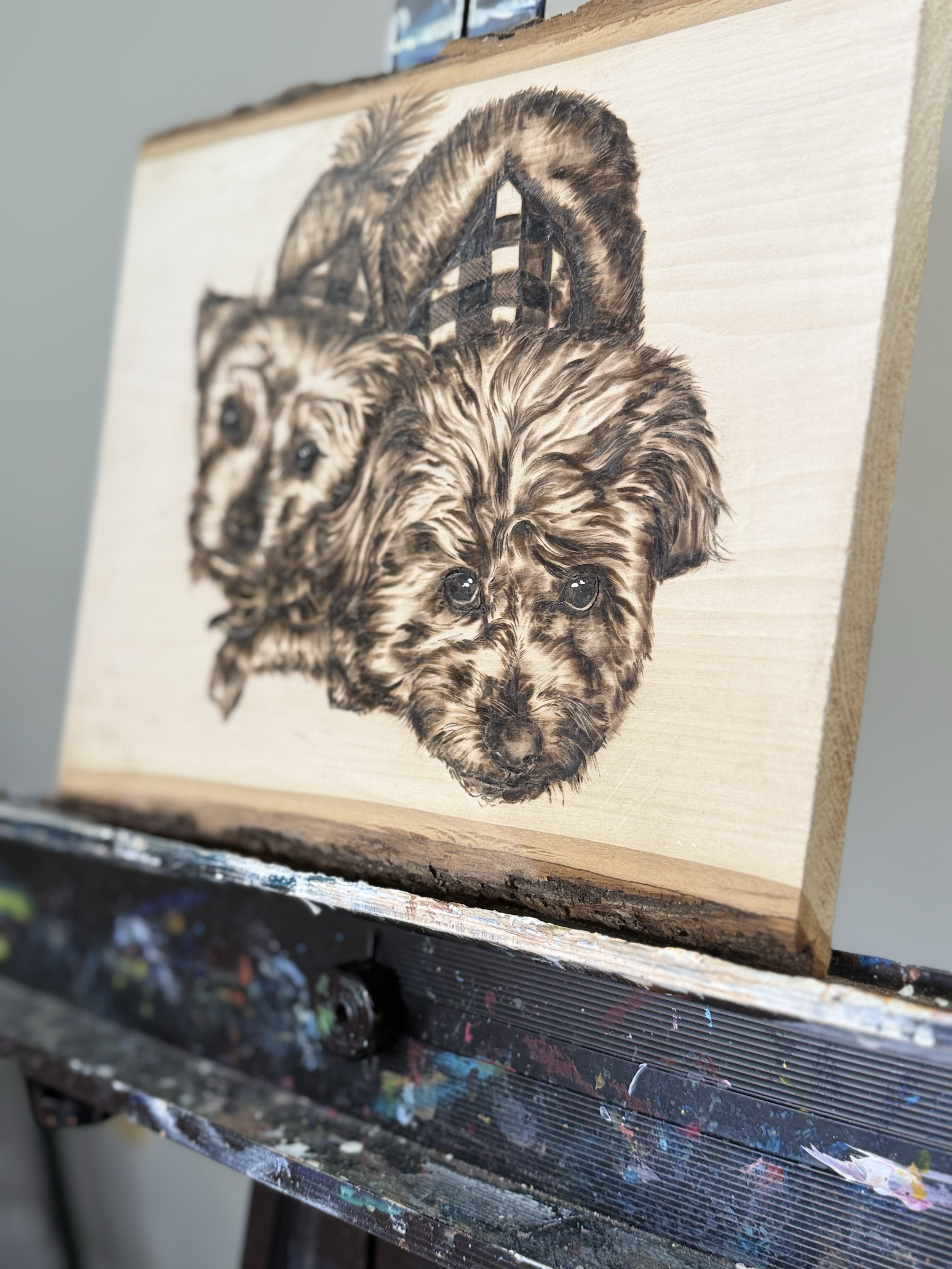 A wood panel painting of two dogs, possibly a terrier and a spaniel, on an artist's easel with paint stains.
