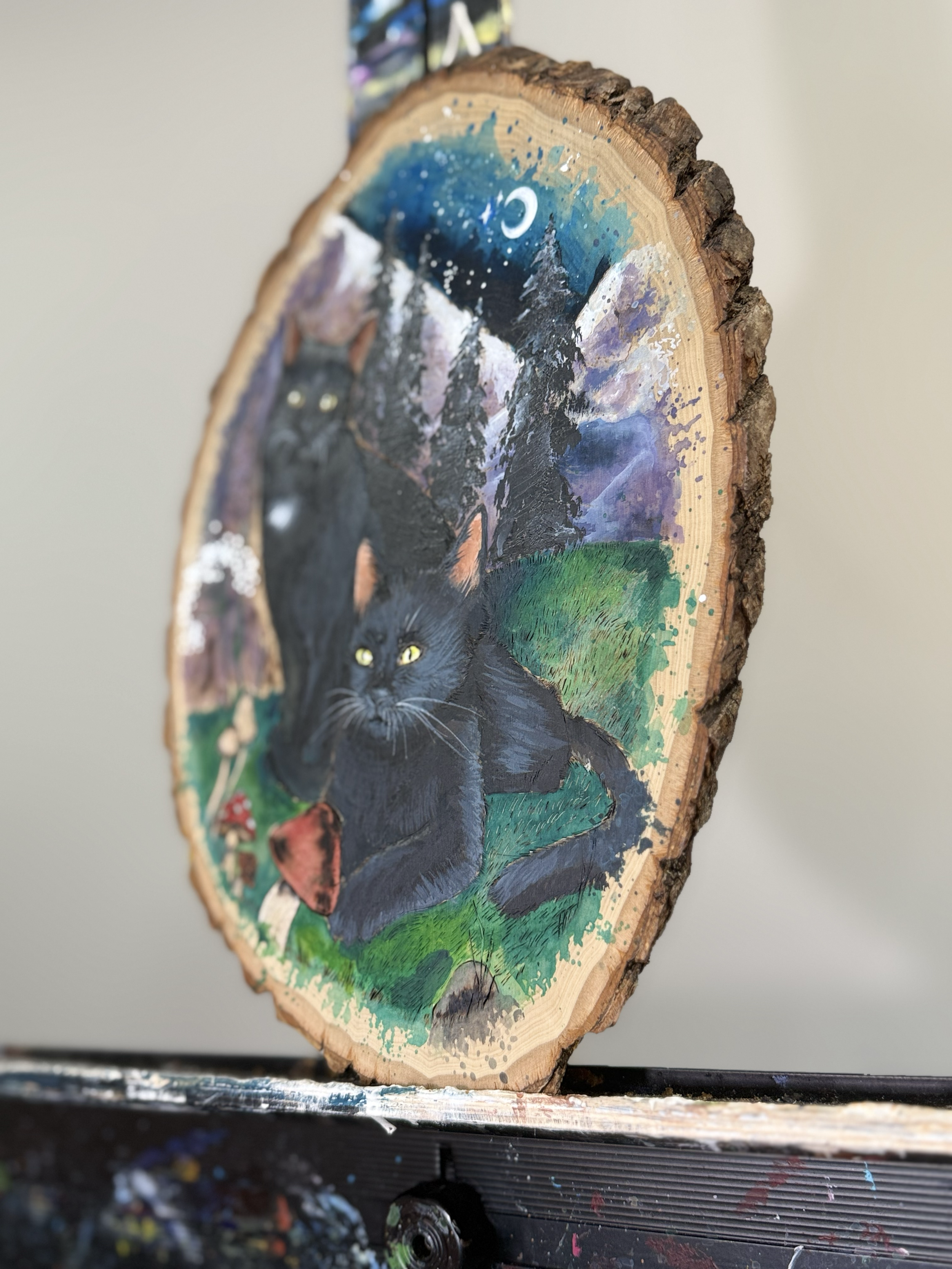 Painted wooden art piece depicting black cats in a wooded, mountain landscape under a night sky with a crescent moon, painted on a cross-section of a tree trunk.