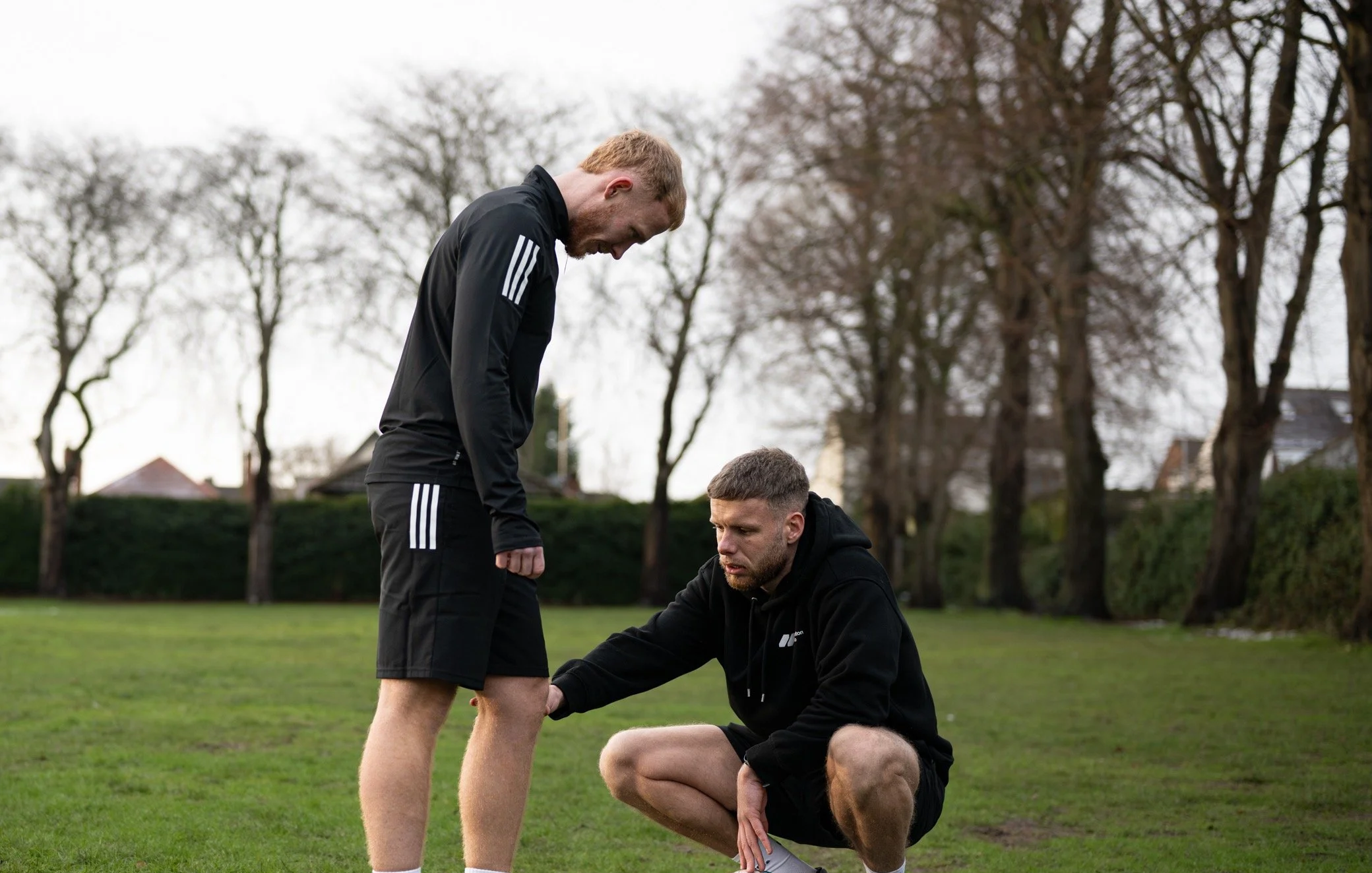 Two men on a grassy field, one kneeling and examining the other's knee, outdoors with trees and suburban houses in the background.