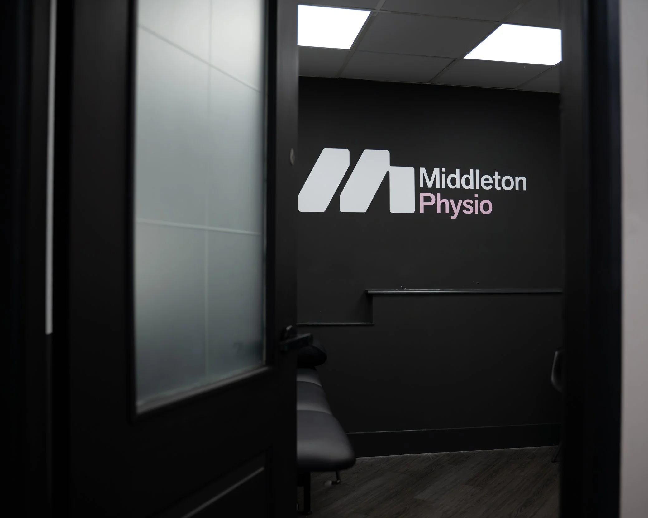 Interior of a clinic room with a black wall, featuring the Middleton Physio logo in white and purple, and a black bench against the wall.