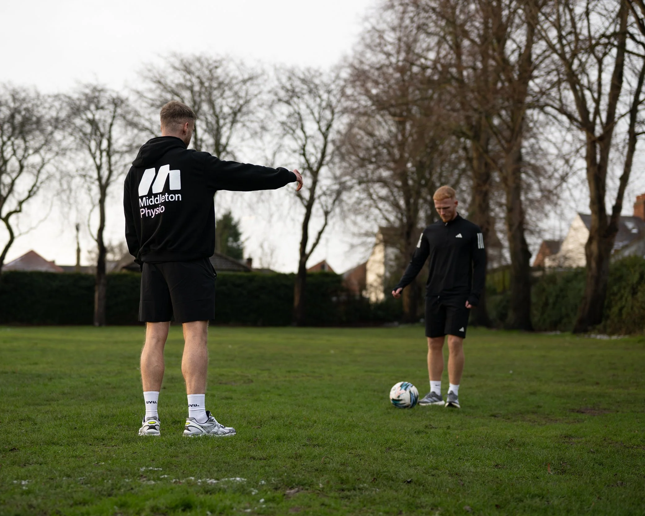 Two men standing on a grassy field, one gesturing towards the other who is preparing to kick a soccer ball, with leafless trees and houses in the background on a cloudy day.