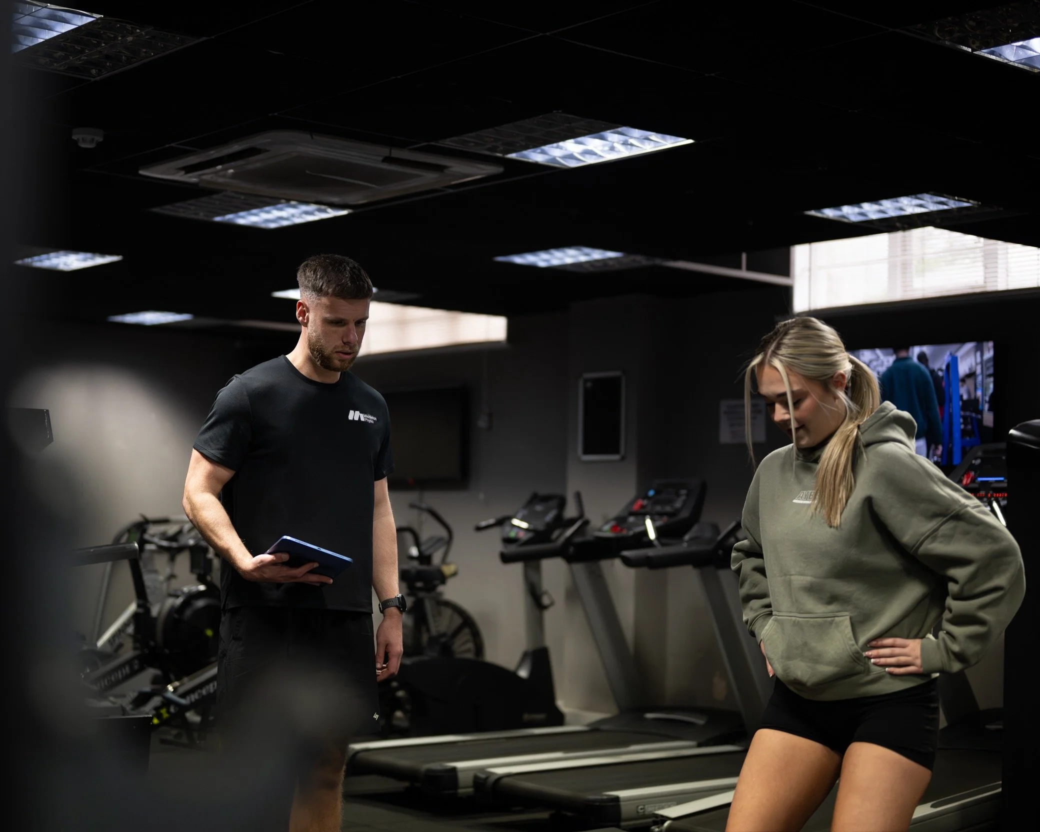 A man and a woman in workout attire inside a gym, with the man holding a clipboard and the woman standing with her hands on her hips near treadmills.